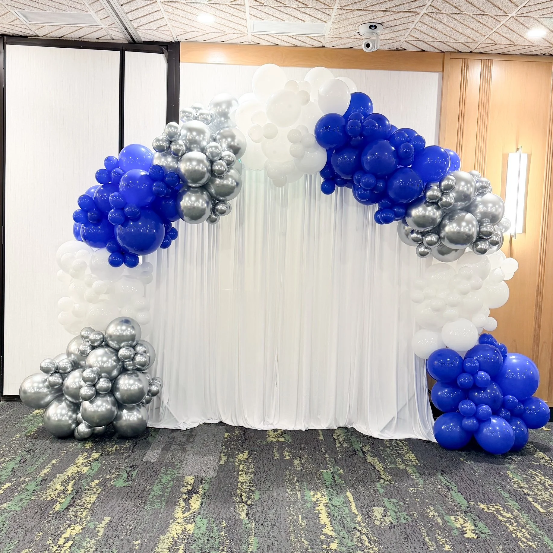 Balloon arch with white, blue, and silver balloons in front of a white curtain backdrop.