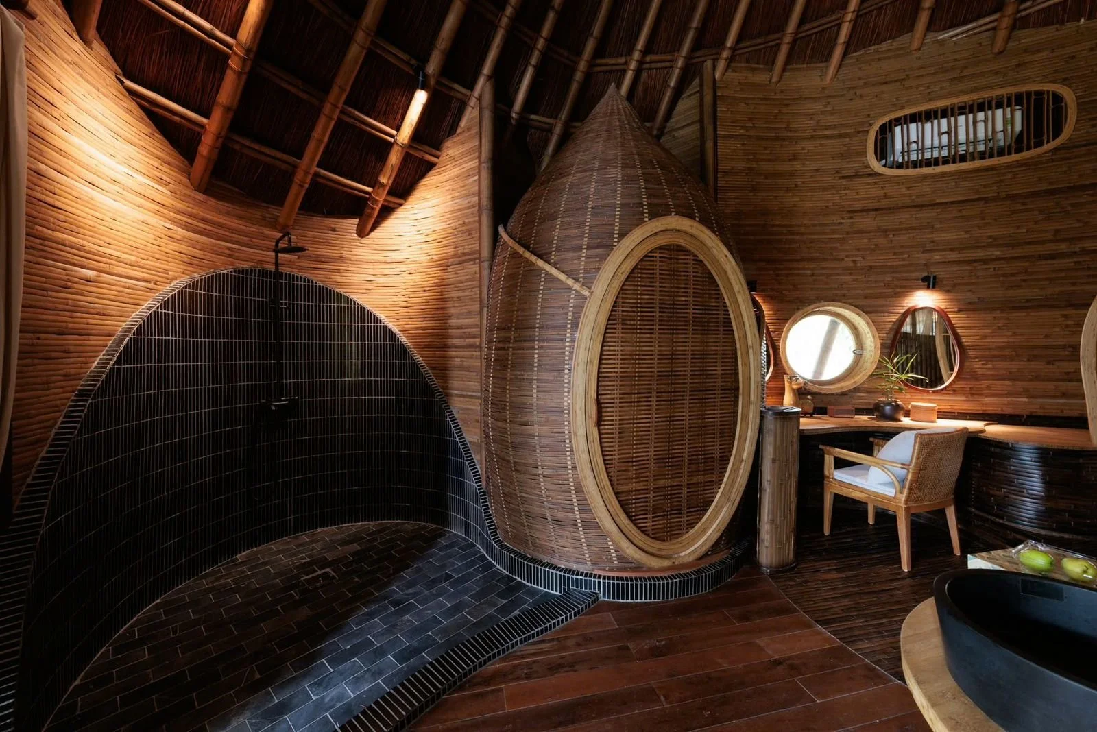 A bathroom with wooden walls and a high, curved thatched ceiling. It features a black-tiled shower with a rounded shape, and a wooden oval-shaped shower enclosure. There is a vanity area with two round mirrors, a wooden countertop, a chair, and some decorative items including a plant and pottery. The floor is dark wood, and there are small windows high on the wall for natural light.