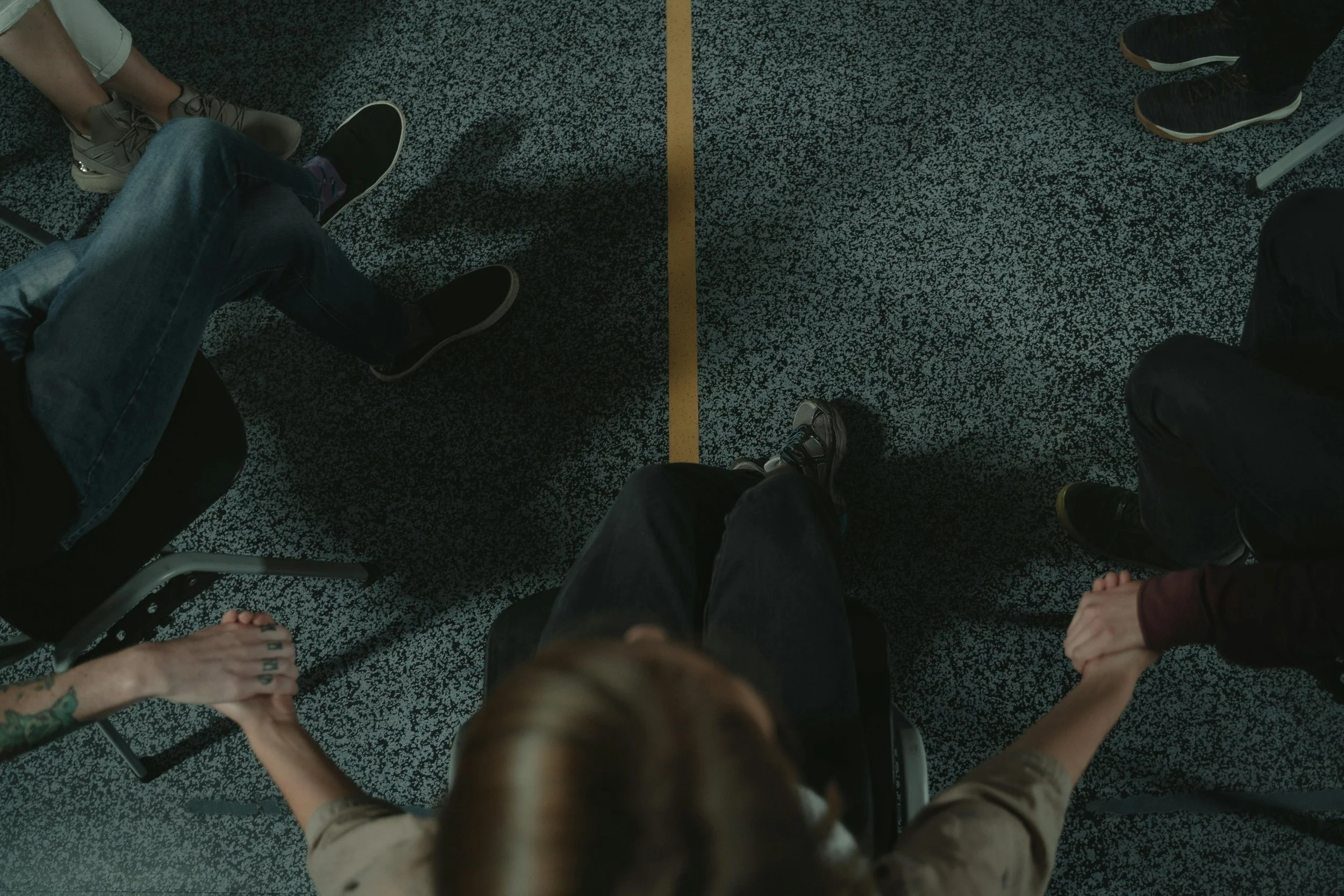 View of a group of people sitting in a circle on a speckled black and white floor, with their legs and feet visible in the image.
