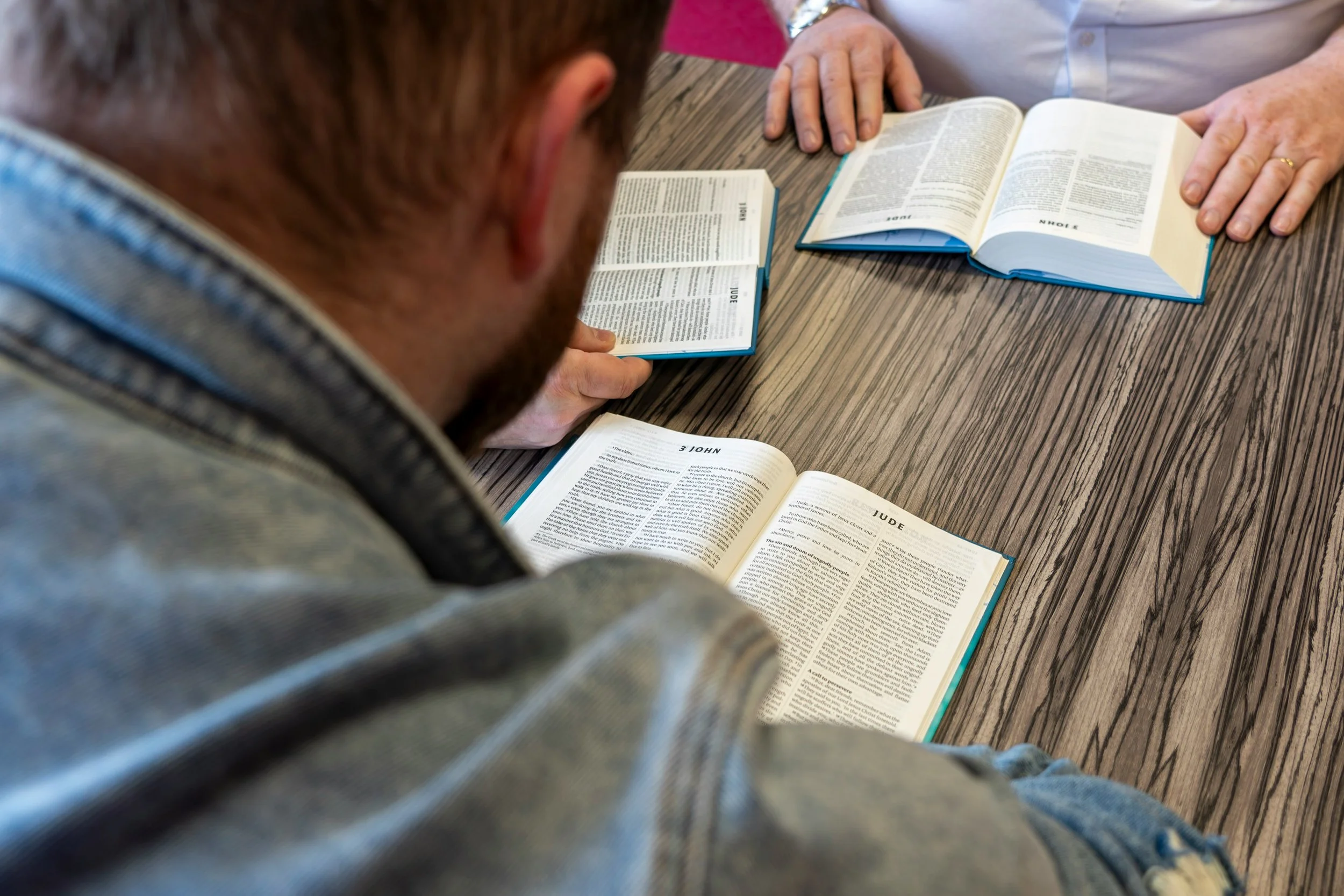 A person in a striped gray shirt sitting at a wooden table with multiple open Bibles in front of them, with at least one person standing nearby.