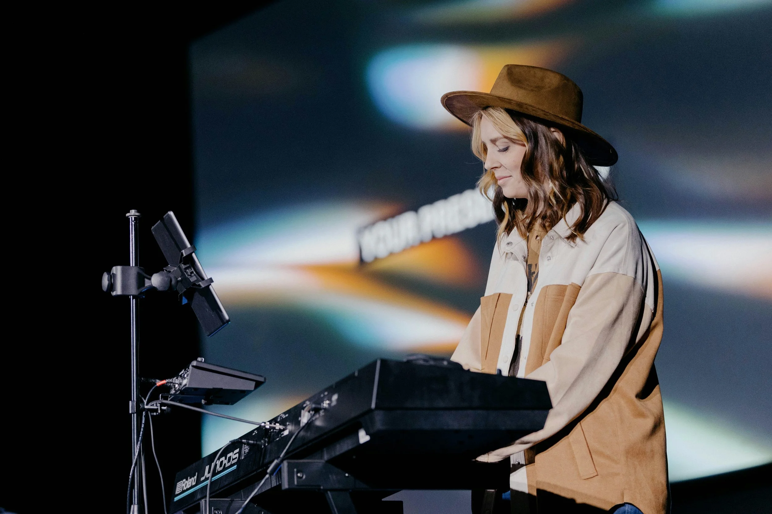 A woman wearing a brown cowboy hat and beige jacket playing a Roland keyboard with a blurred colorful background.