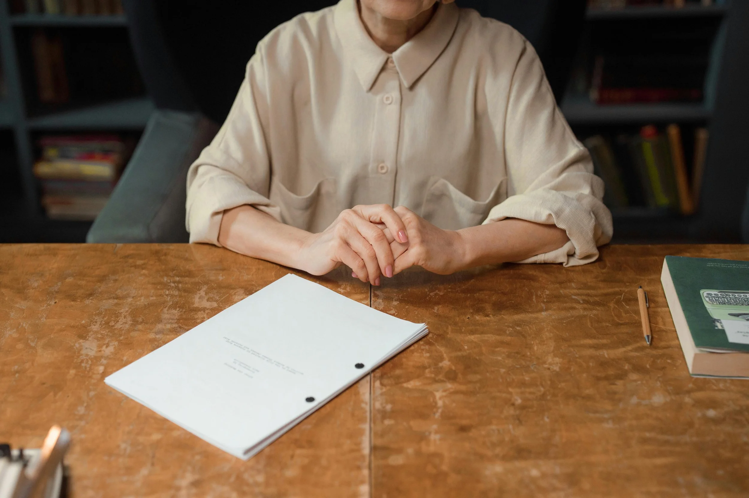 A person wearing a cream-colored button-up shirt sitting at a wooden table with papers, a book, a pen, and a calculator.