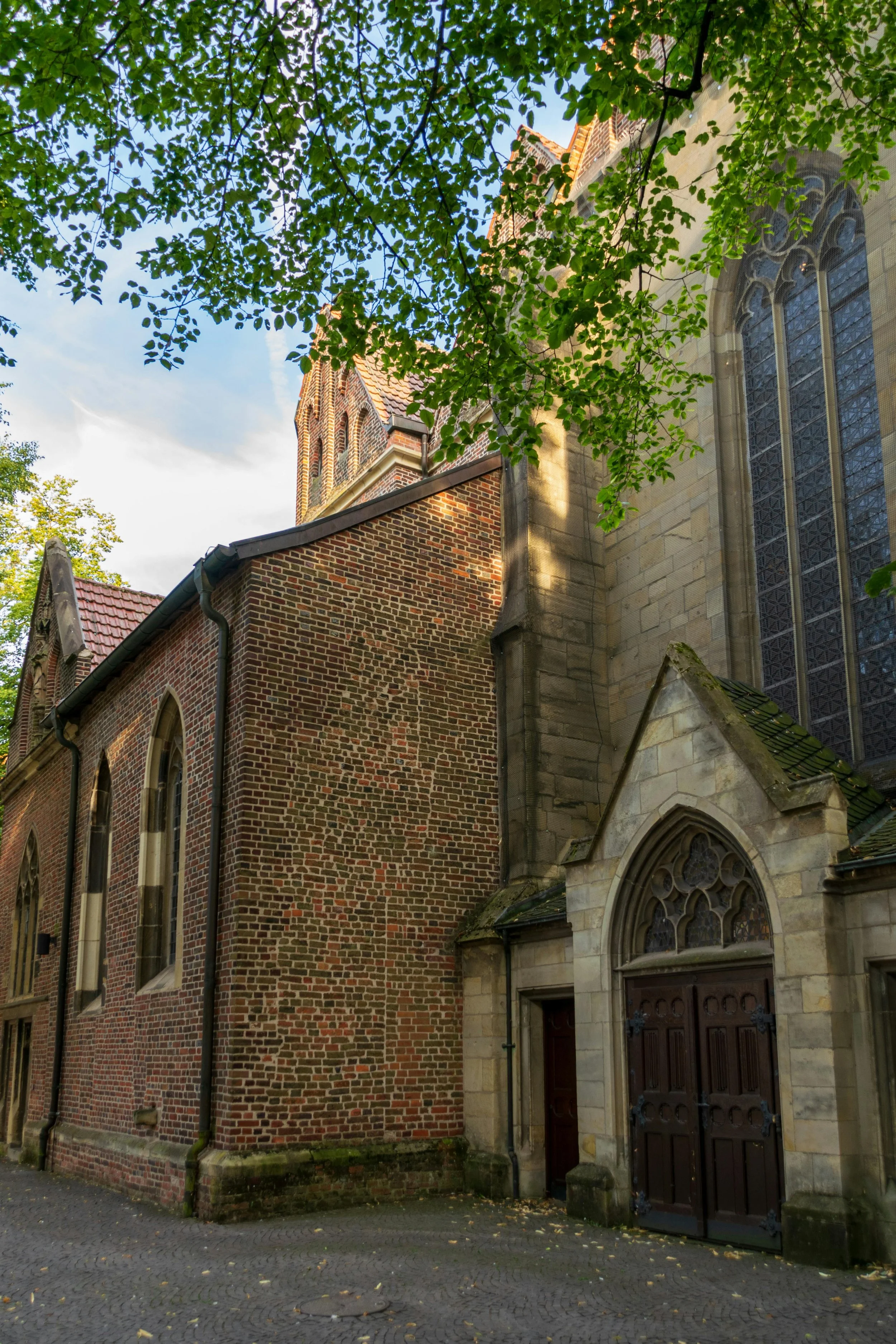 View of an old brick church with large stained glass windows and an arched wooden door, partially obscured by green tree branches.