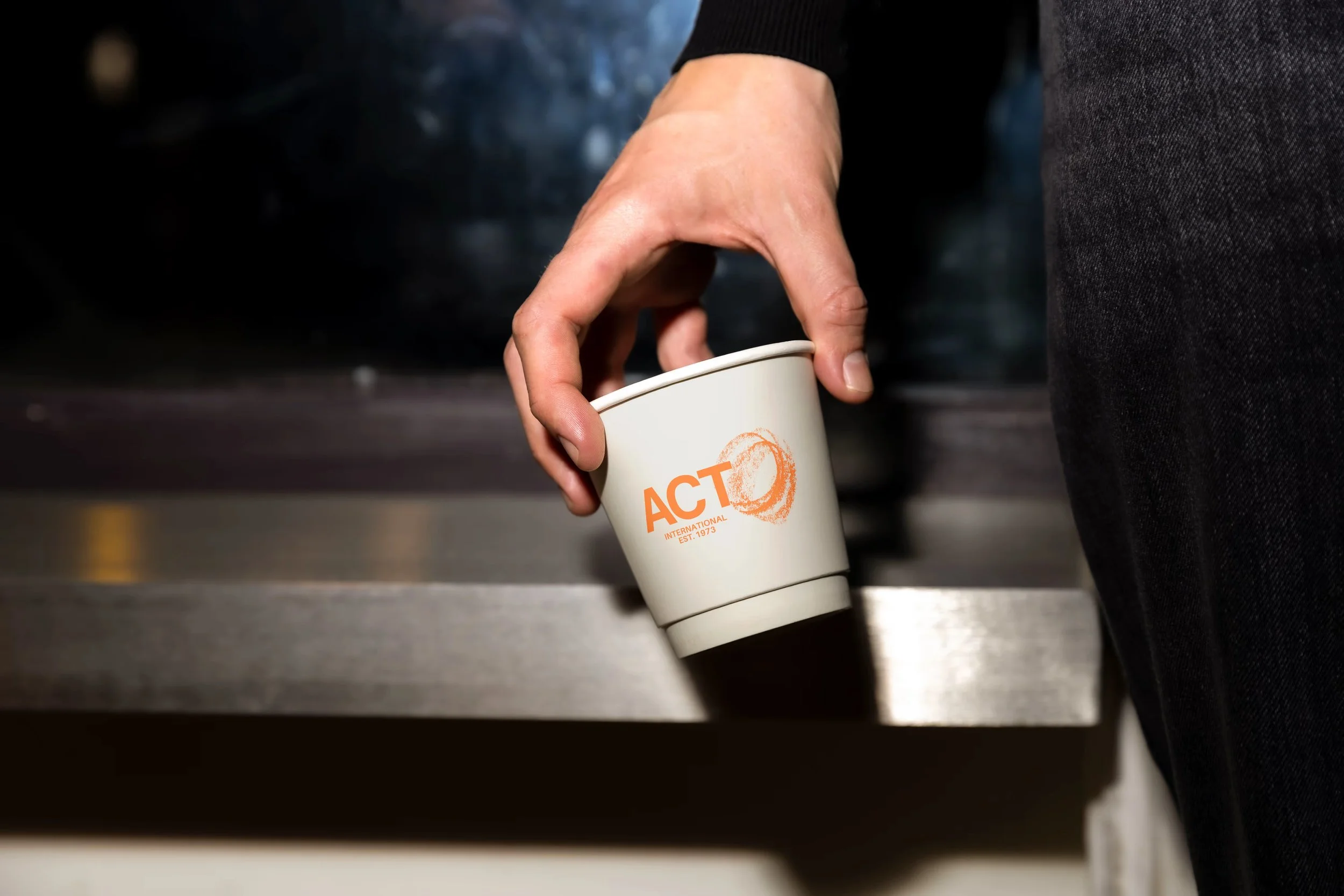 A person wearing black clothing holding a white disposable cup with orange text that reads 'ACT' and 'INTERNATIONAL' and a circular logo, near a trash or recycling bin.