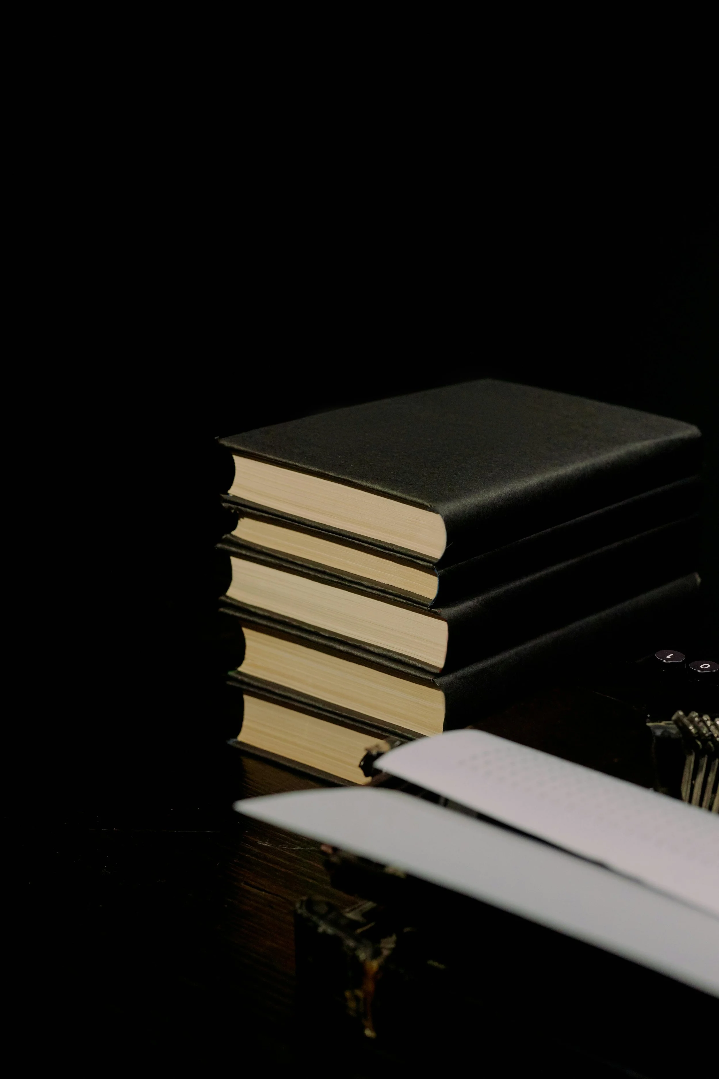 Stacked black notebooks or journals on a dark surface, with a pen or mechanical pencil and a notebook or paper nearby.