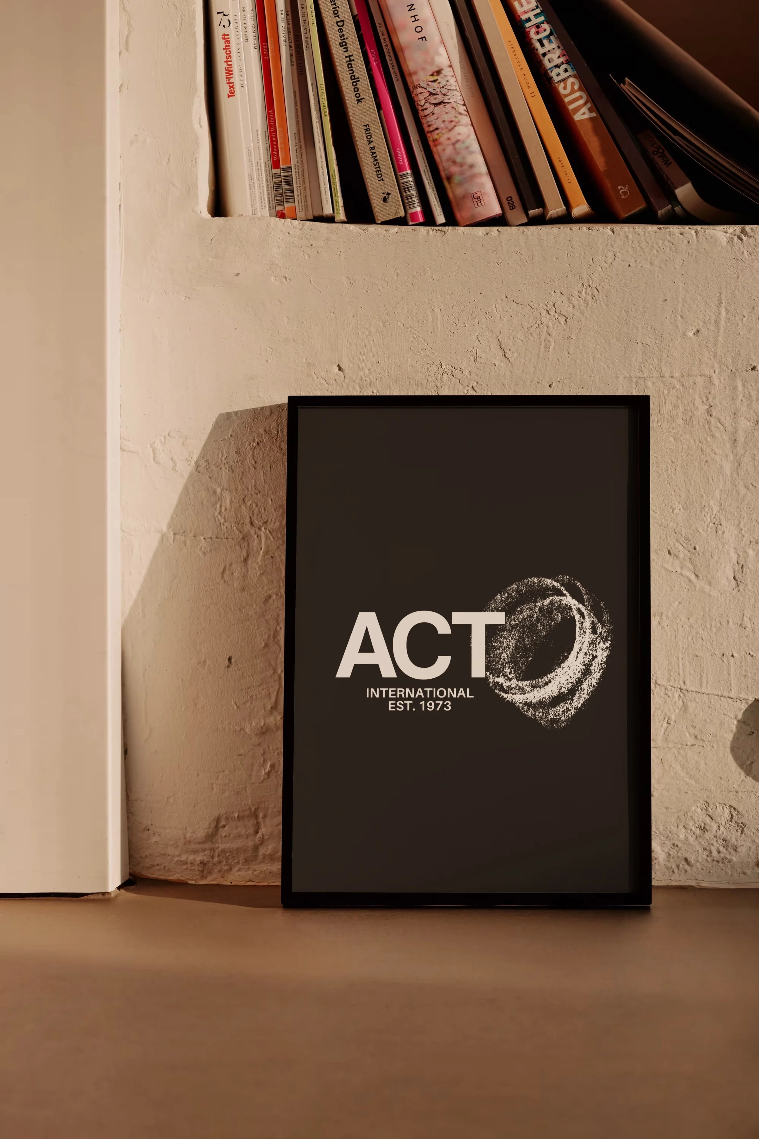 Close-up of a shelf with books and a framed poster. The books include titles on design, art, and literature, positioned vertically on the shelf. The framed poster displays the word 'ACT' with a circular design and the text 'INTERNATIONAL' and 'EST. 1973' beneath it. The background features a textured beige wall.