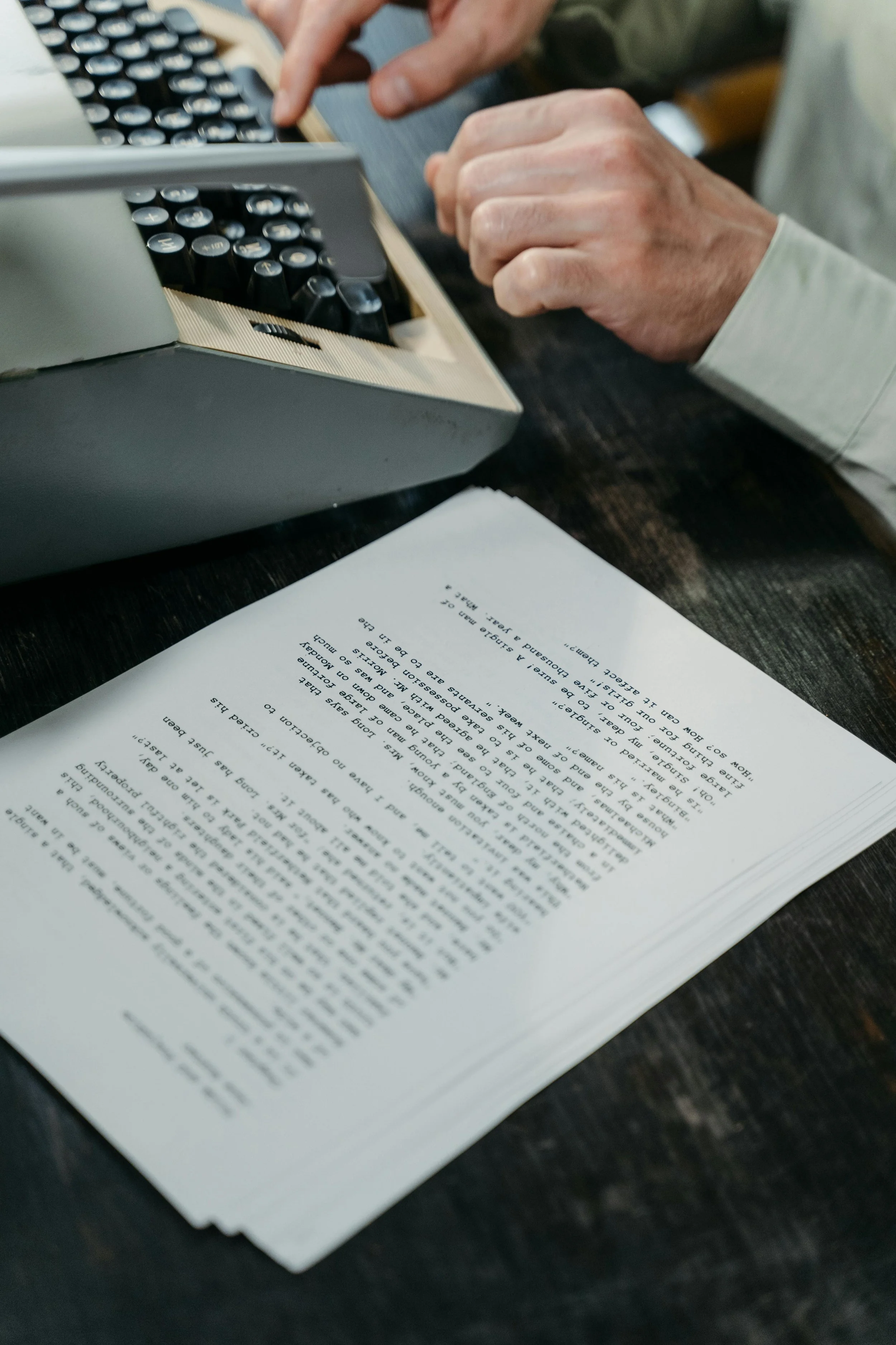 A person on a vintage typewriter while reading a printed document on a dark wooden table.