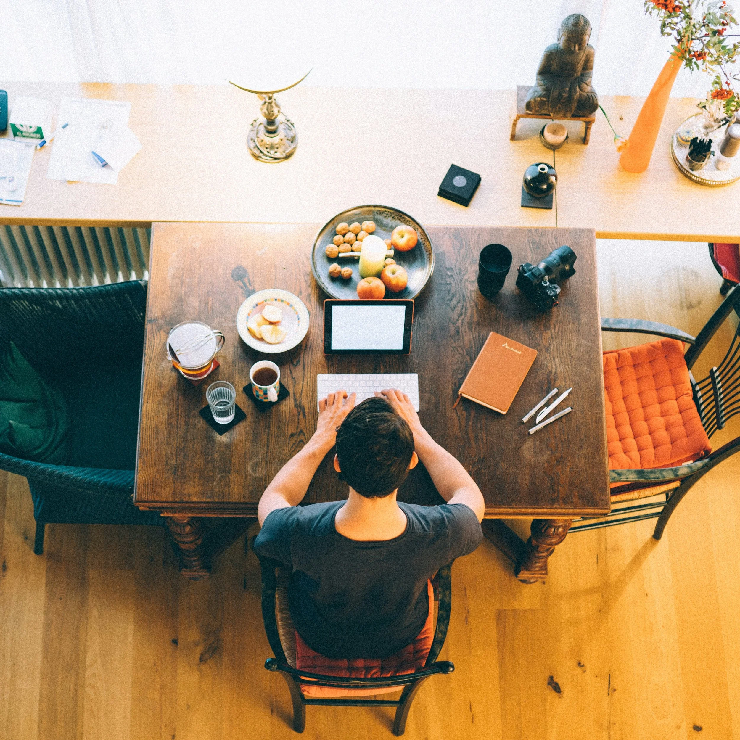 Overhead view of a person sitting at a wooden dining table with various items including fruits, notebooks, a camera, and decorative objects, in a brightly lit room with wooden flooring.