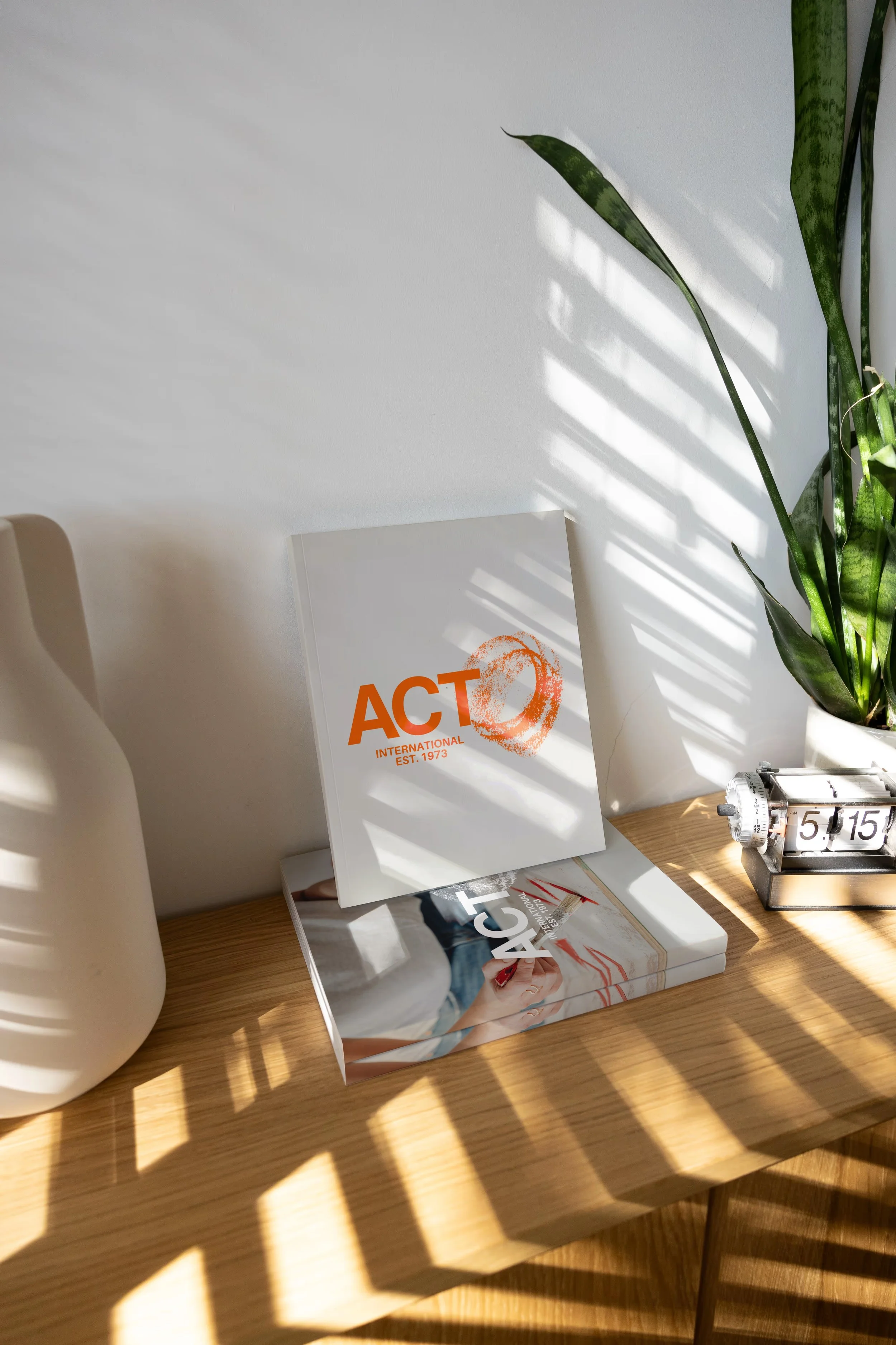 A desk with various items: a white mug, a book titled 'ACT International,' a magazine, a white box, a potted plant with long green leaves, and a clock showing 5:15. Sunlight casts shadows on the desk.