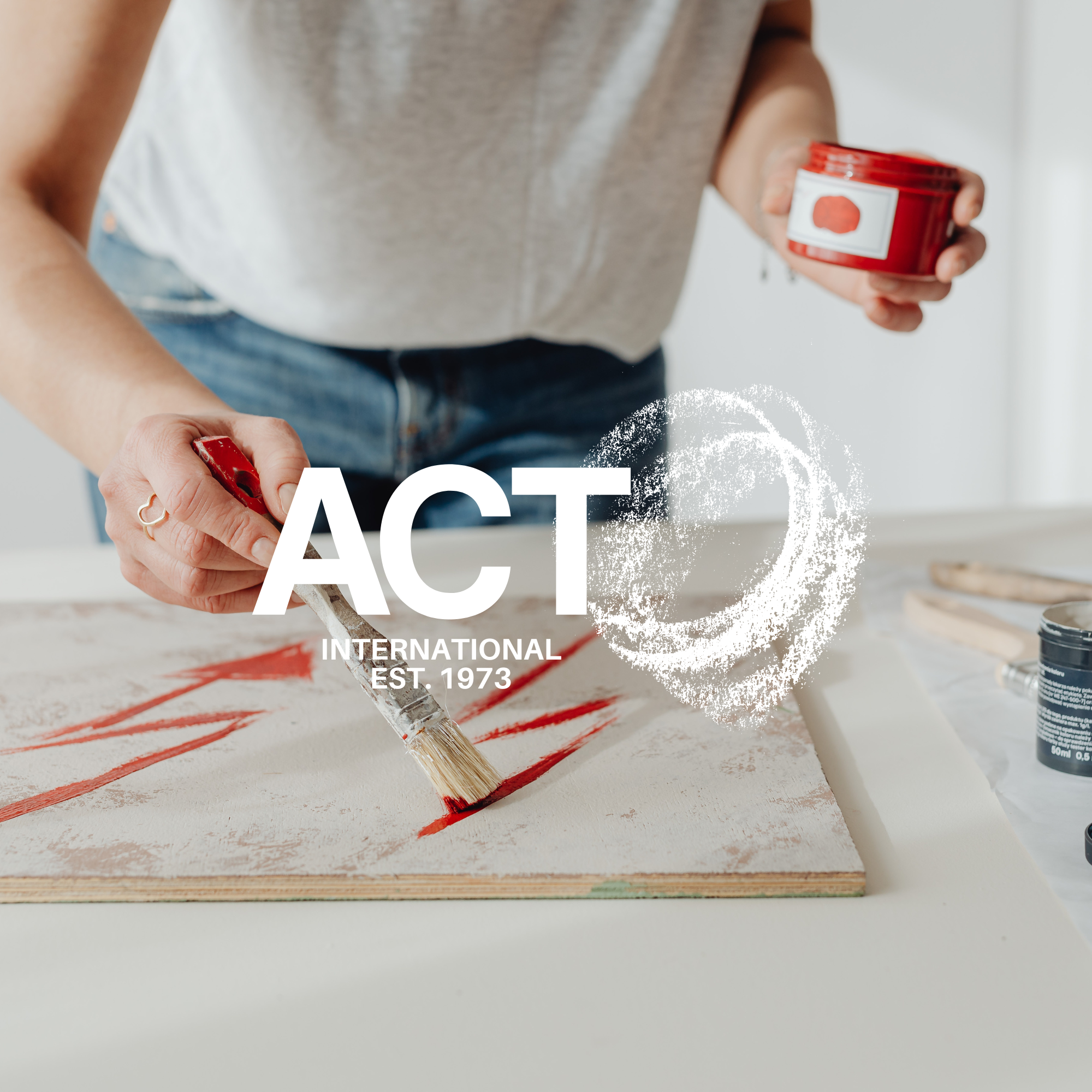 Person brushing red paint on a piece of wood, with cans of paint nearby. Part of a white shirt and blue jeans visible.