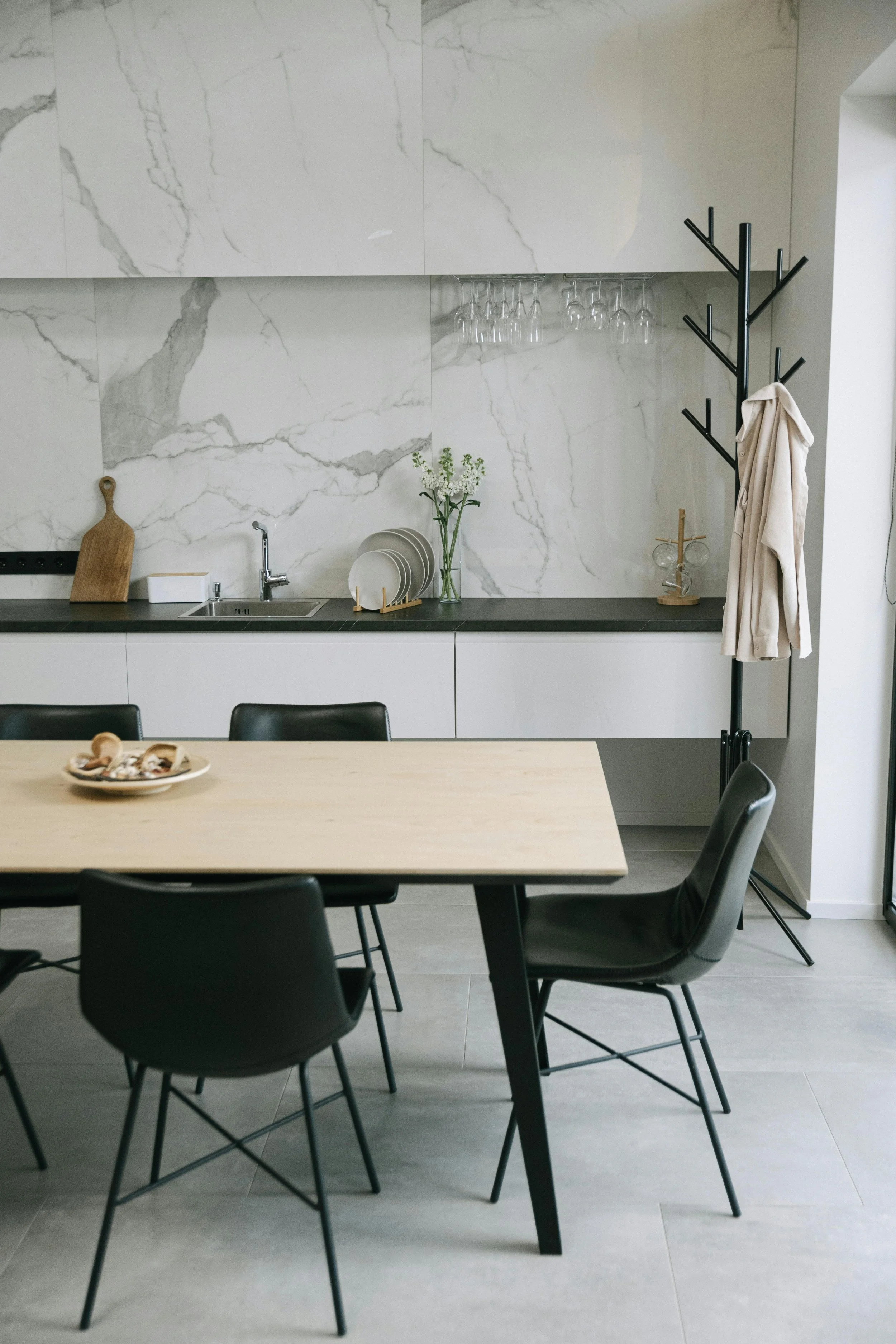 Modern kitchen with marble backsplash, black countertop, dish rack with dishes, flowers in a vase, wine glasses hanging, wooden cutting board, and black coat rack with a beige coat. Dining table with black chairs in foreground.
