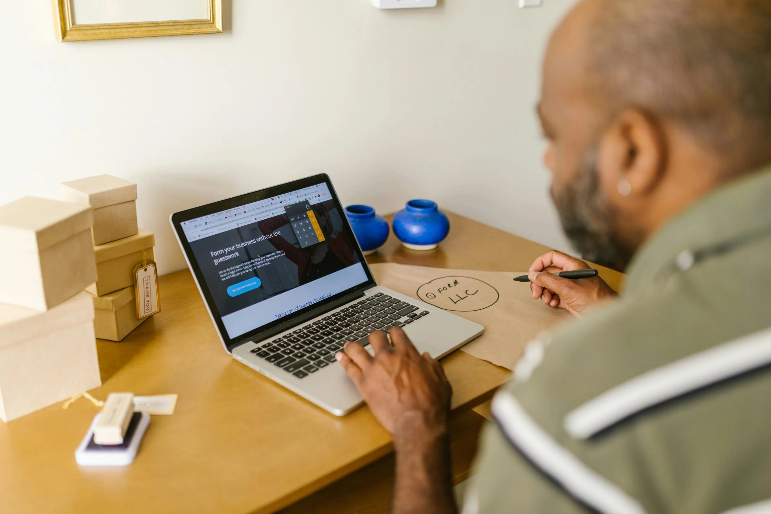 Man working on a laptop at a desk with boxes, a notepad, and decorative blue vases in a home office.