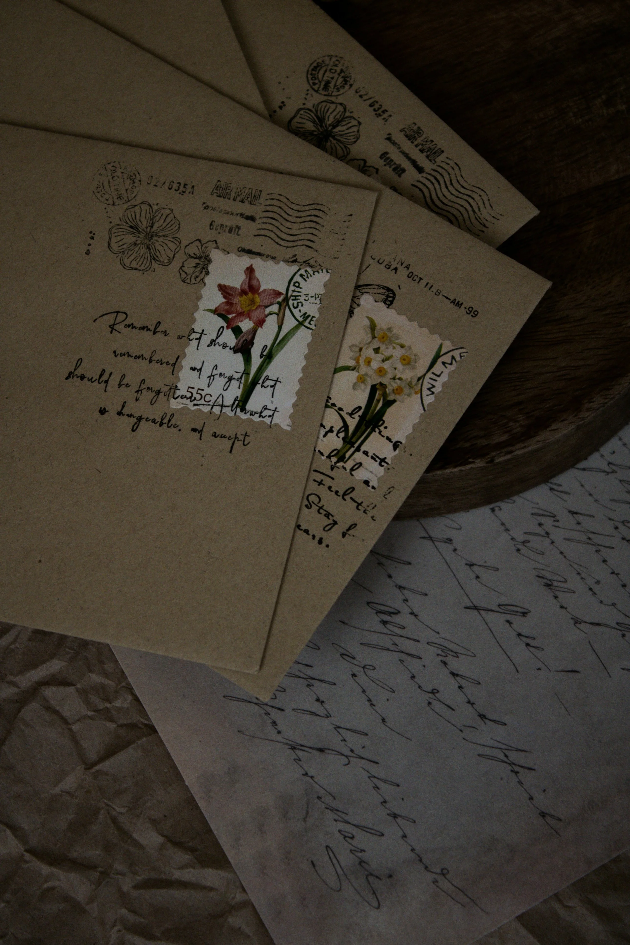 A close-up of vintage-style postal letters with handwritten text, floral stamps, and postage markings, placed on crumpled brown paper and wood surfaces.