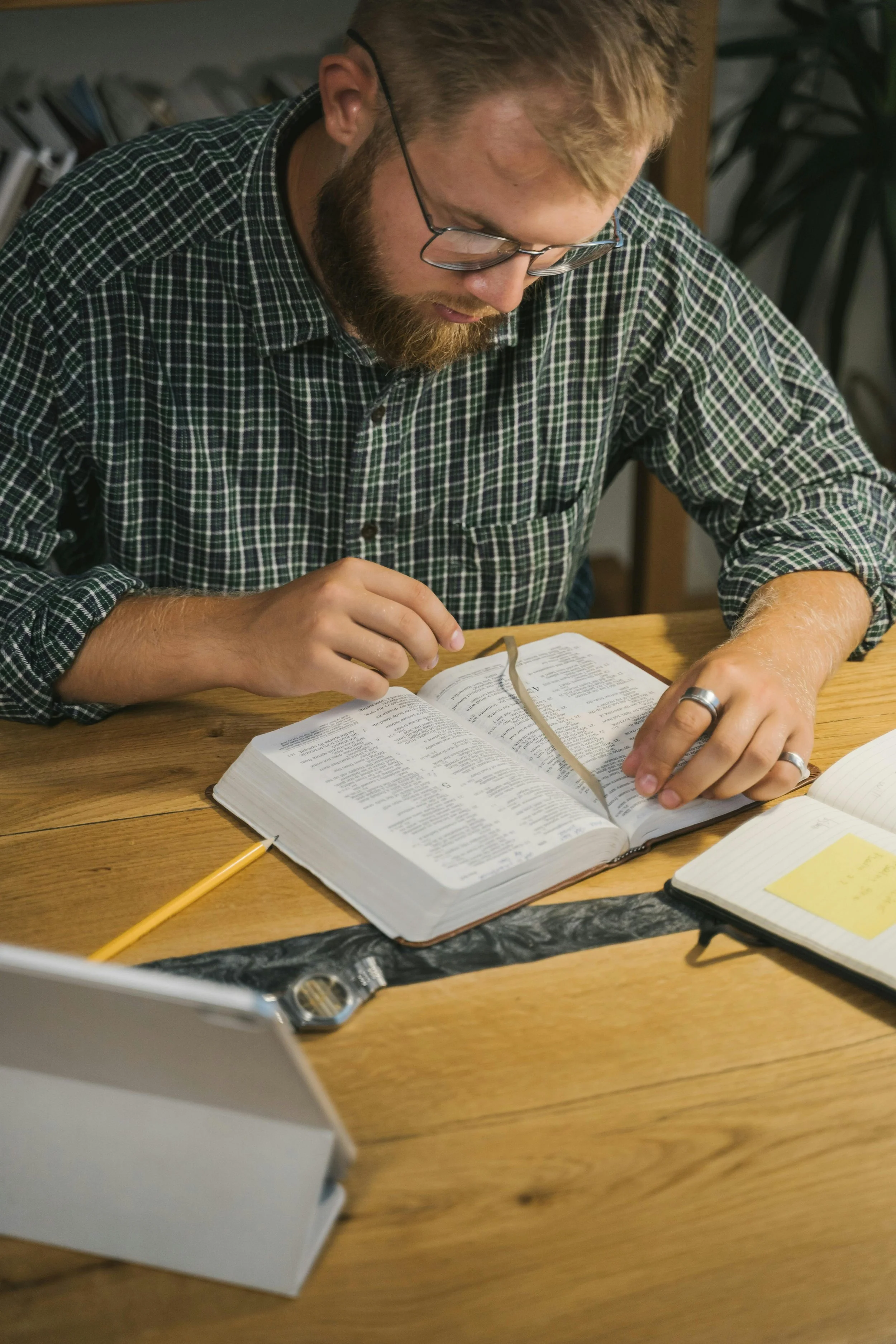 A man with glasses and a beard wearing a green checkered shirt reading a large book at a wooden table.