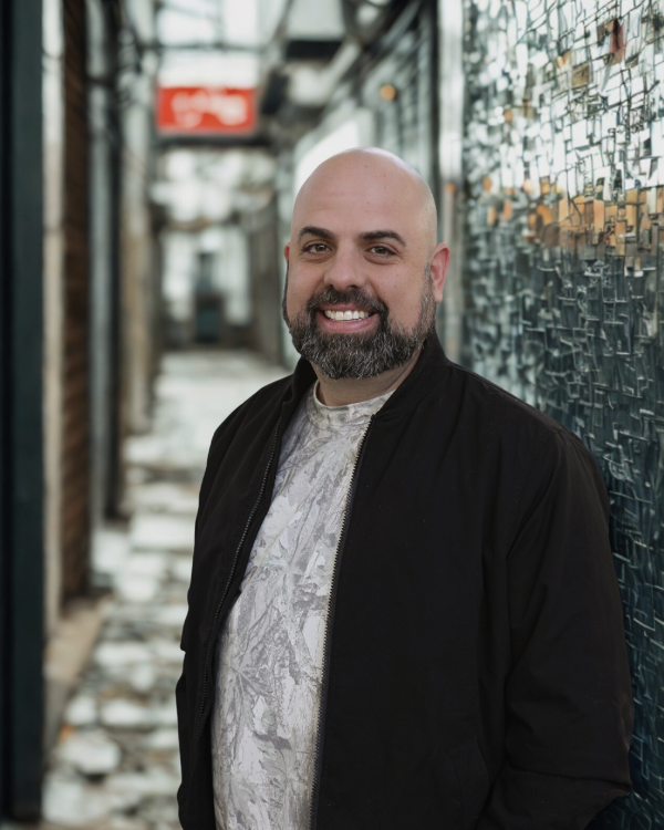 Smiling man with a beard and bald head leaning against a decorated glass wall.