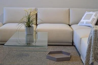 A modern white sectional sofa with a geometric patterned pillow, a glass coffee table with a potted plant, and a gray hexagonal tray on a gray carpet in a minimalist living room.