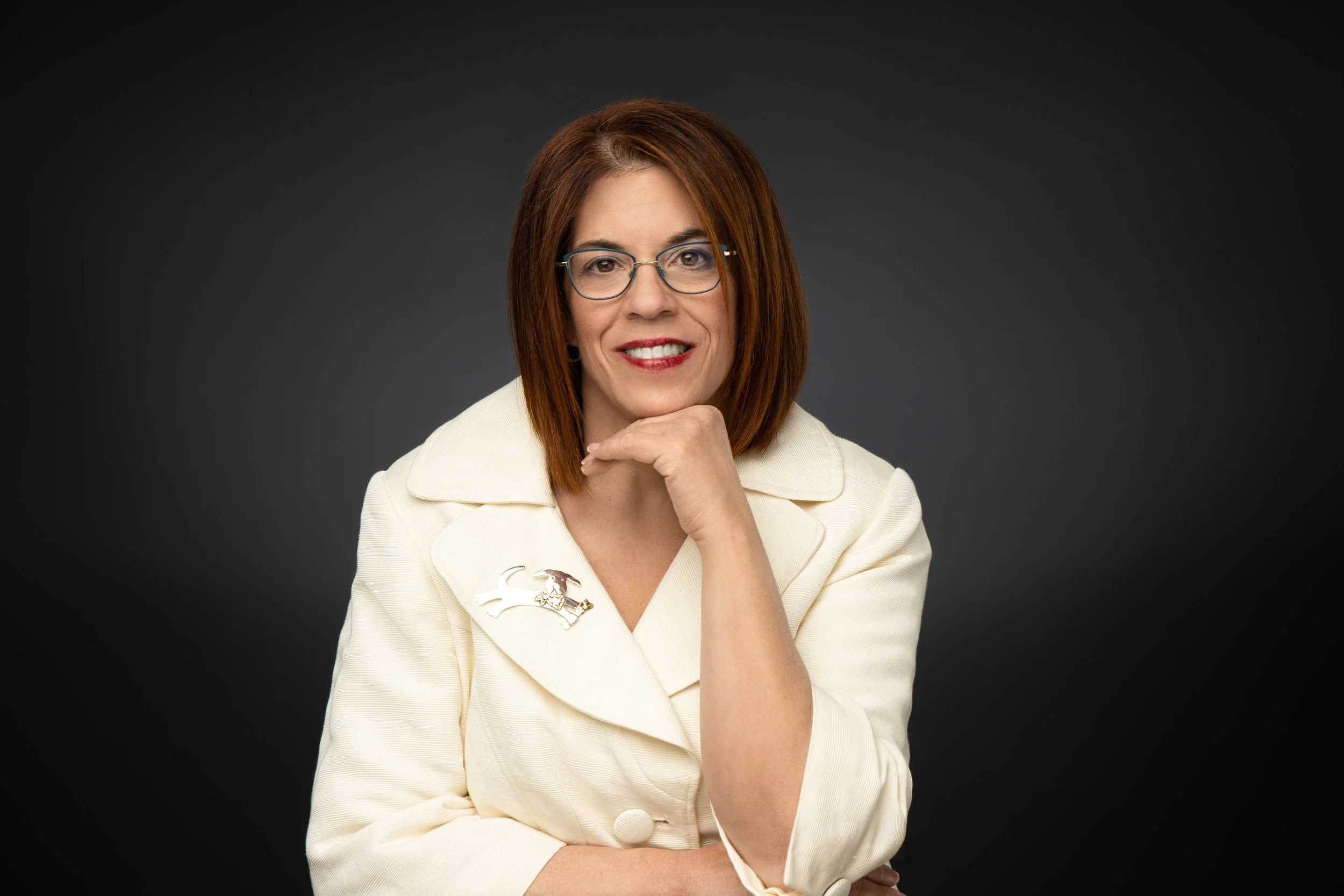 A woman with shoulder-length red hair and glasses, wearing a cream-colored blazer with a dog-shaped brooch, smiling and resting her chin on her hand against a dark gray background.
