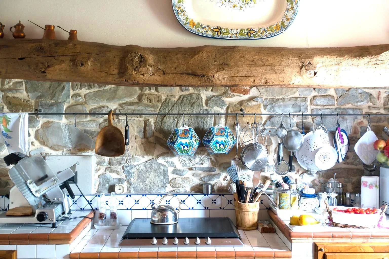 A rustic kitchen with a stone and wood wall, hanging pots, pans, and utensils, a stovetop with a kettle, a utensil holder, a basket of tomatoes, and a dresser with kitchen supplies.