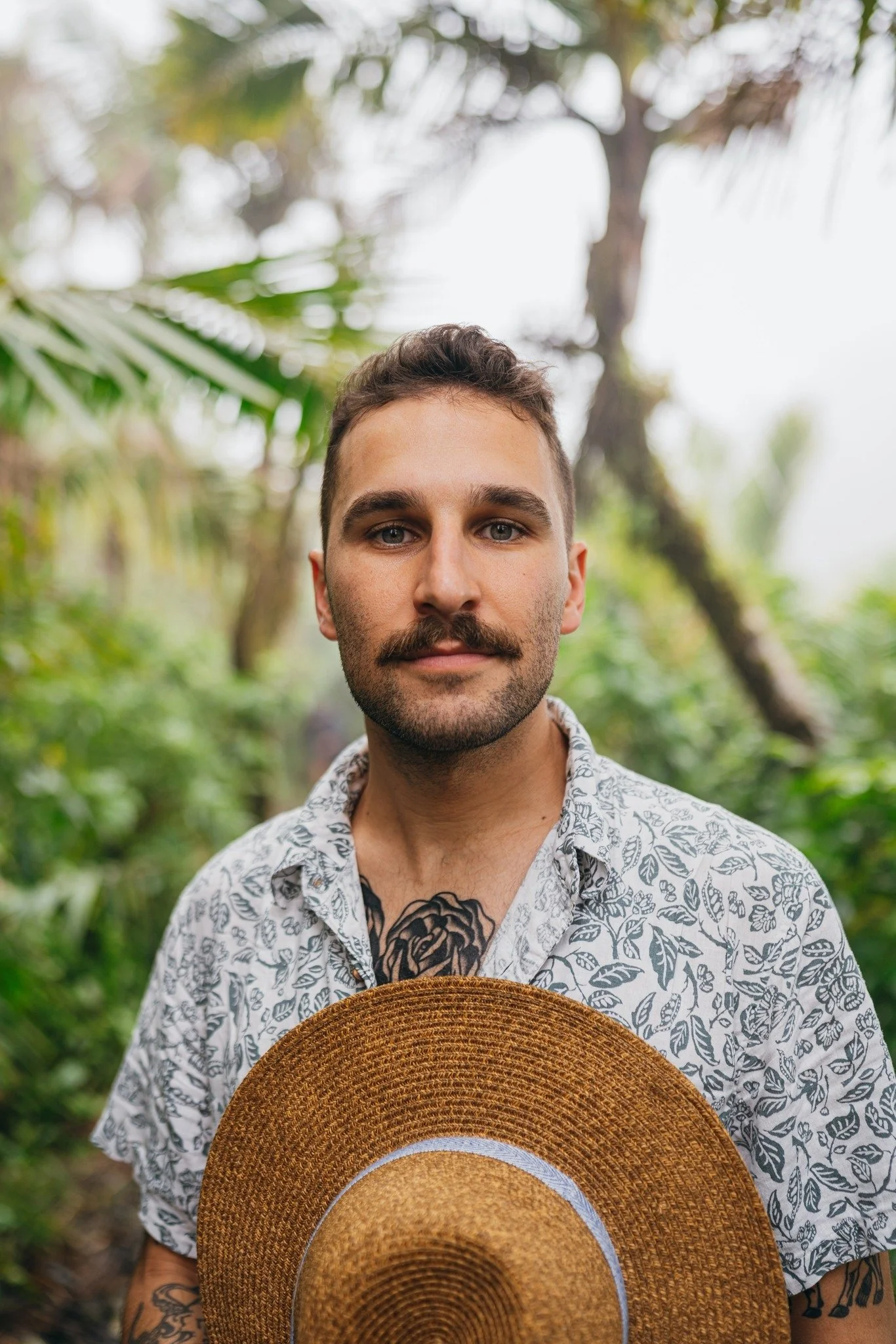 A young man with short dark hair and a beard holding a large straw hat, standing outdoors in a lush green tropical setting with blurred trees and foliage in the background.