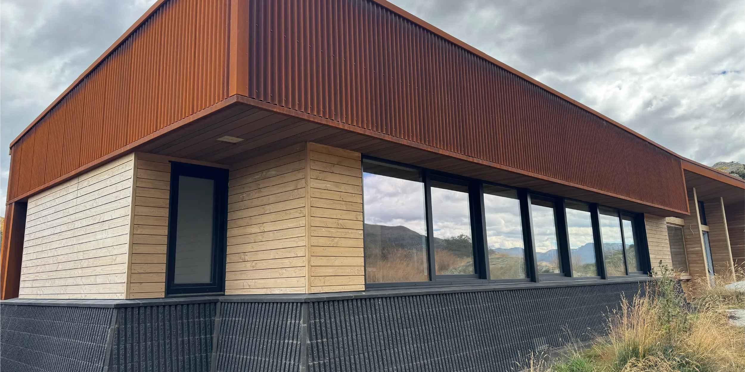 Modern house with a wooden exterior and large windows, set against a cloudy sky with mountains in the background.