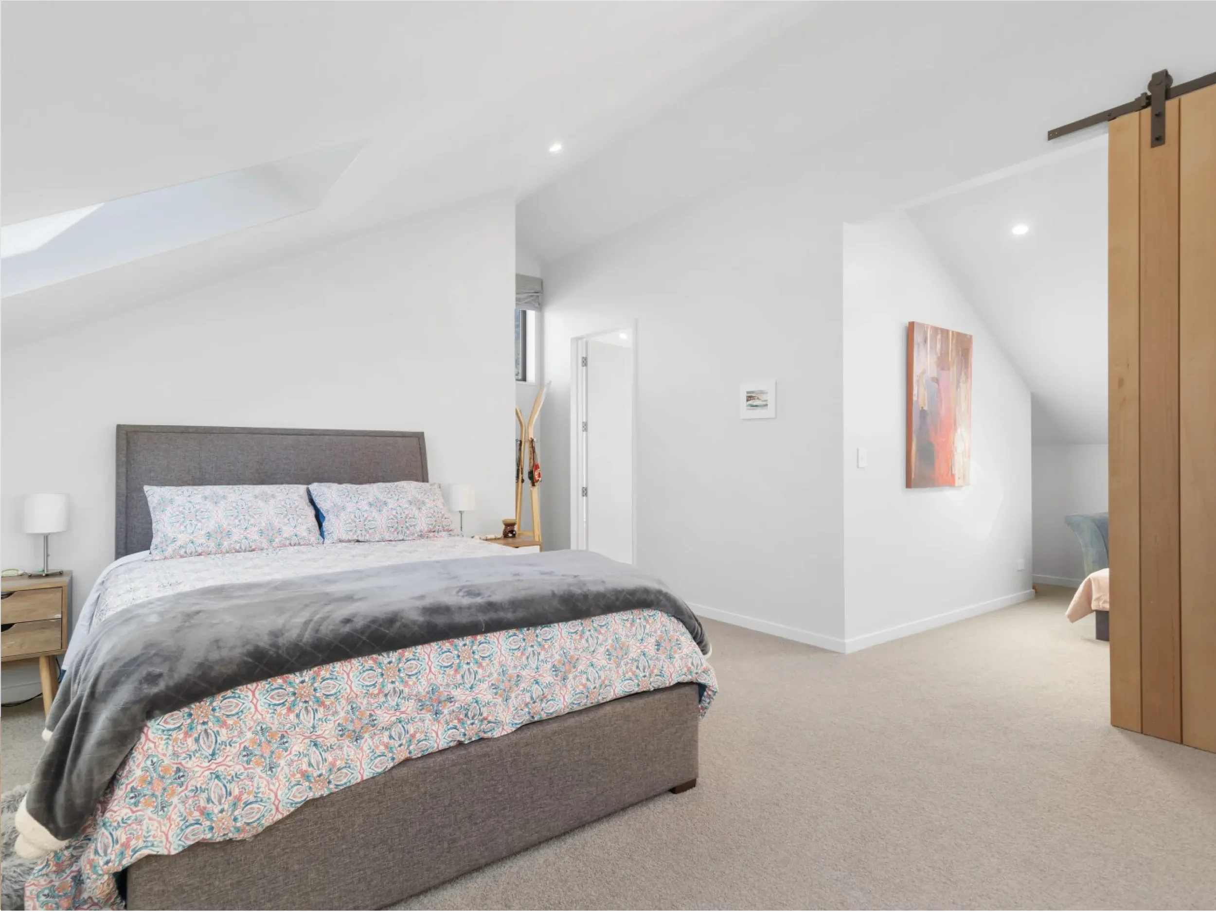 A bedroom with a bed that has a gray upholstered headboard and patterned bedding, two white bedside lamps, a small window, and a wooden sliding barn door.