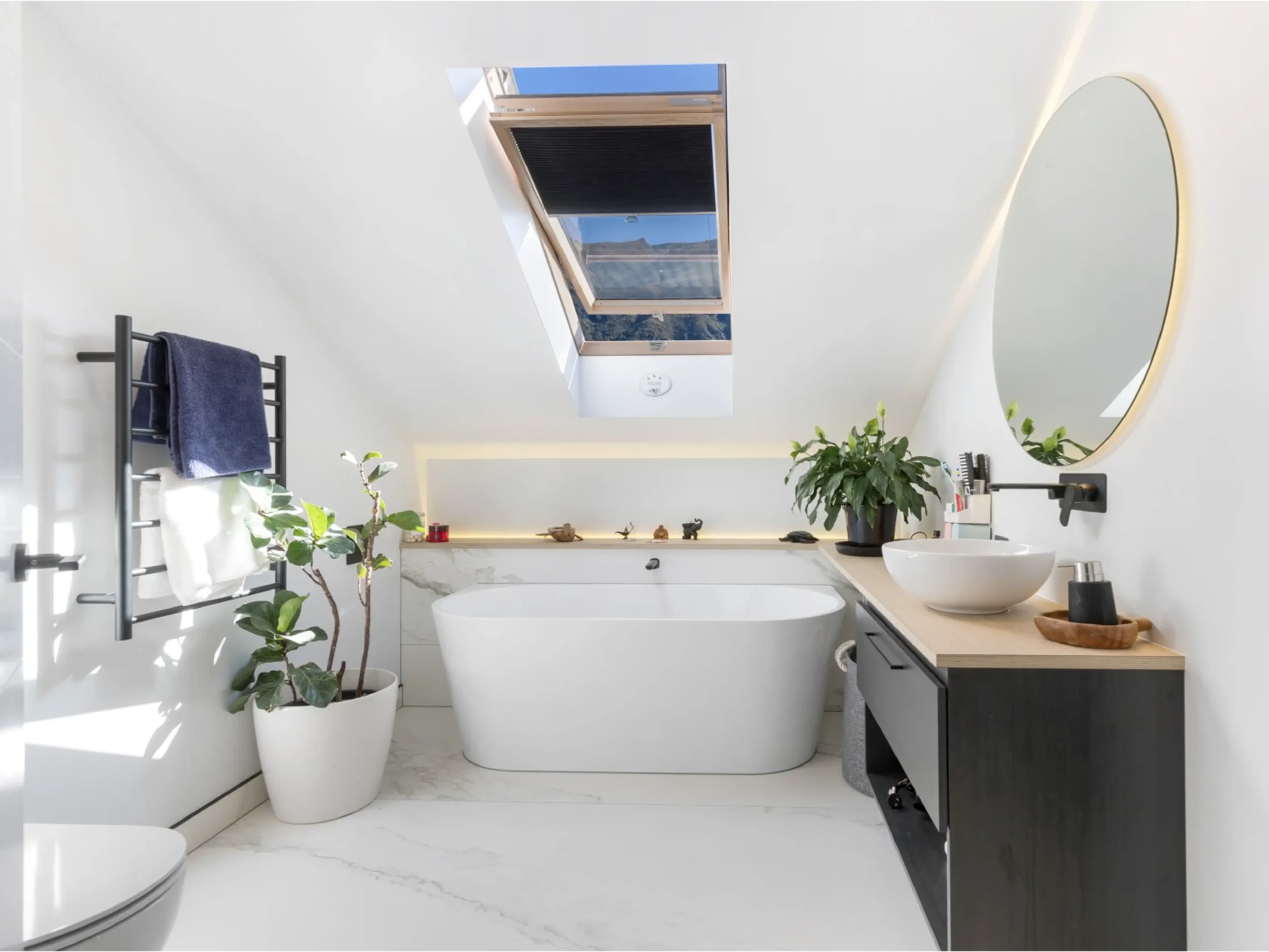 Modern bathroom featuring a freestanding bathtub beneath a skylight window, a large round mirror on the wall, a black towel rack with a towel, potted plants, and minimalist decor with natural light.