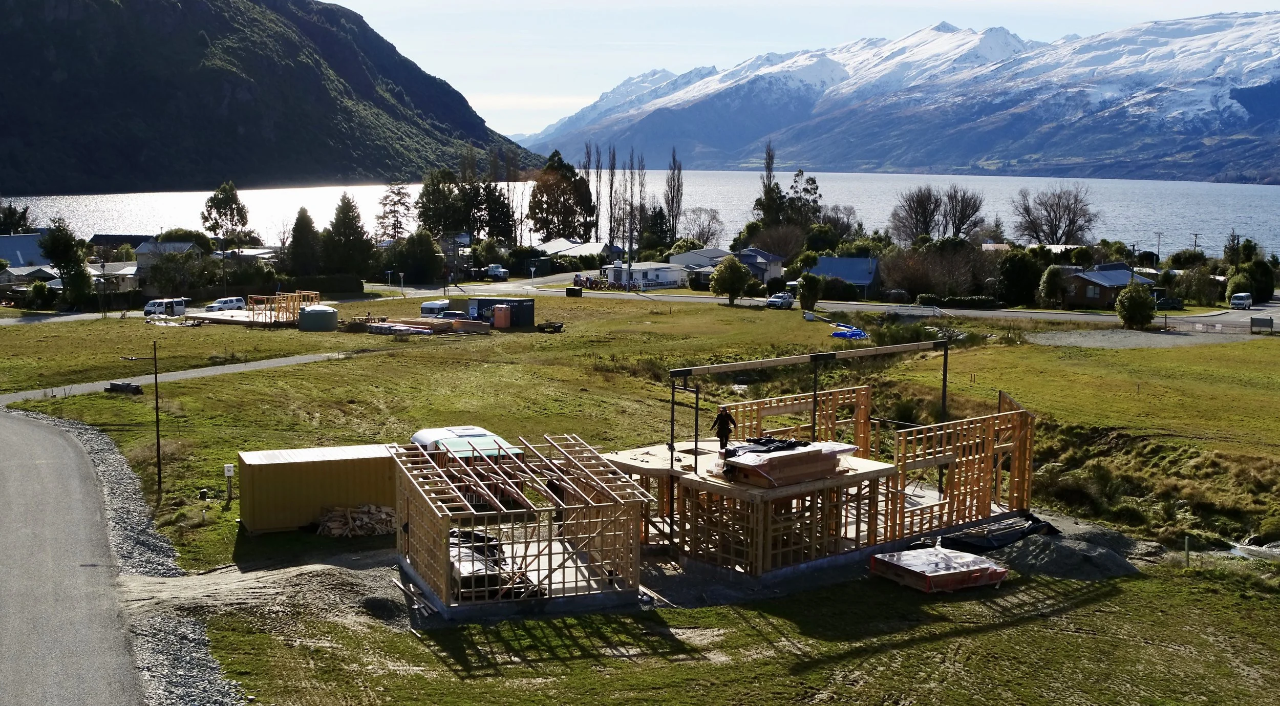 A scenic view of a lake surrounded by snow-capped mountains and lush green hills, with a small residential area and a construction site in the foreground.