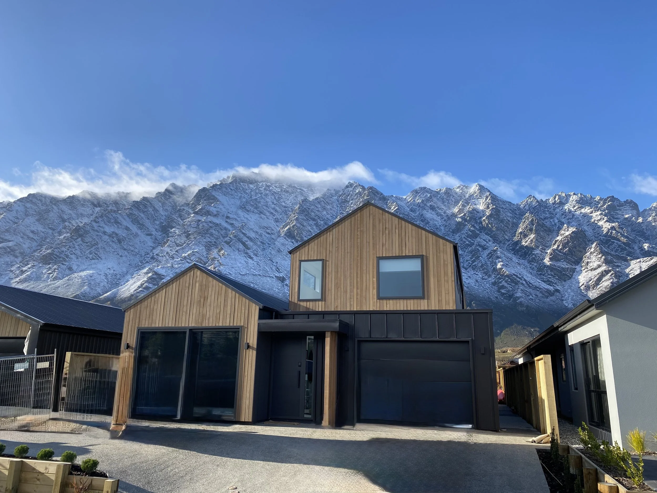 Modern wooden and black houses in front of snow-capped mountains under a clear blue sky.