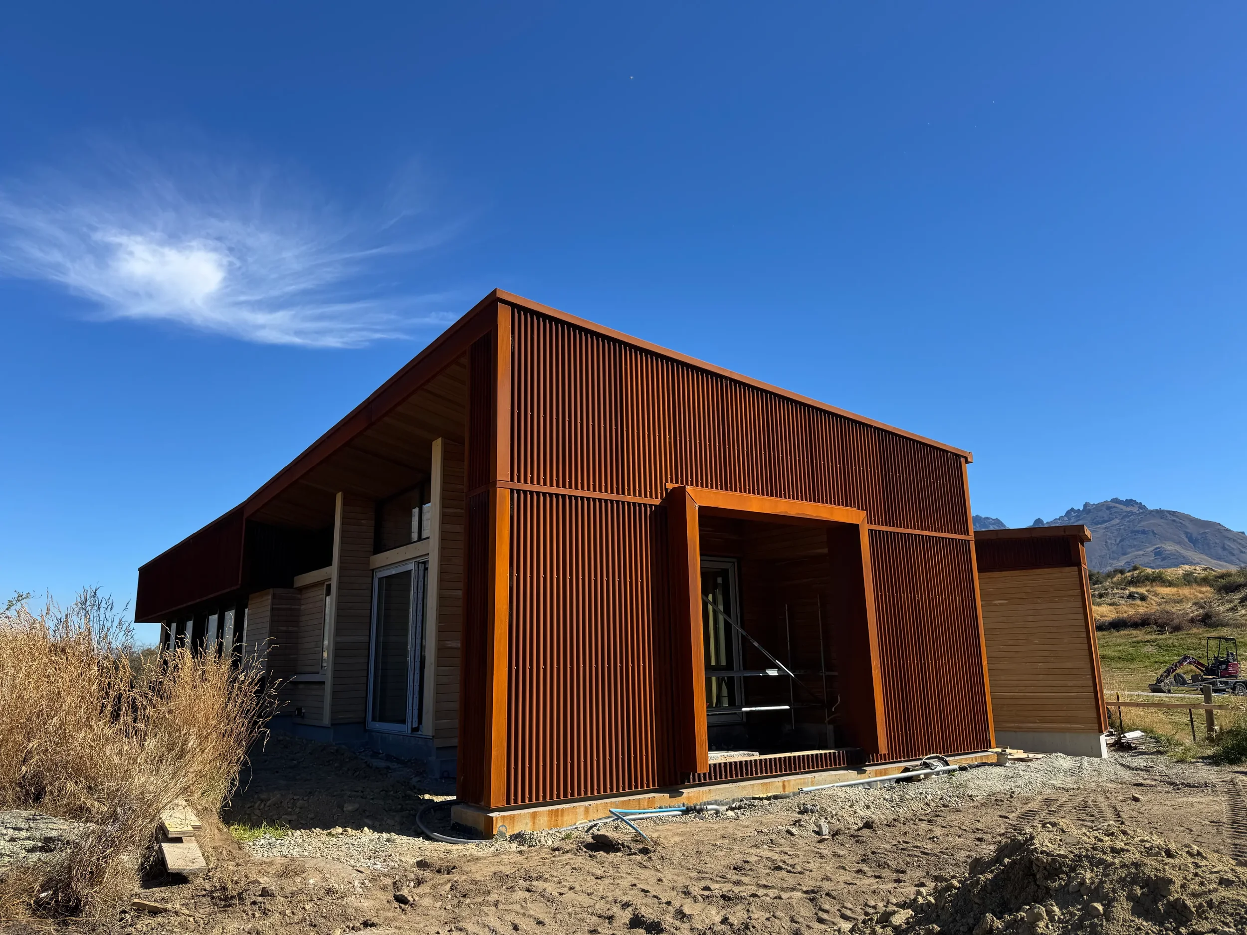 A modern wooden building under construction with vertical slats on the outside, set against a clear blue sky with a few clouds and distant mountains in the background.