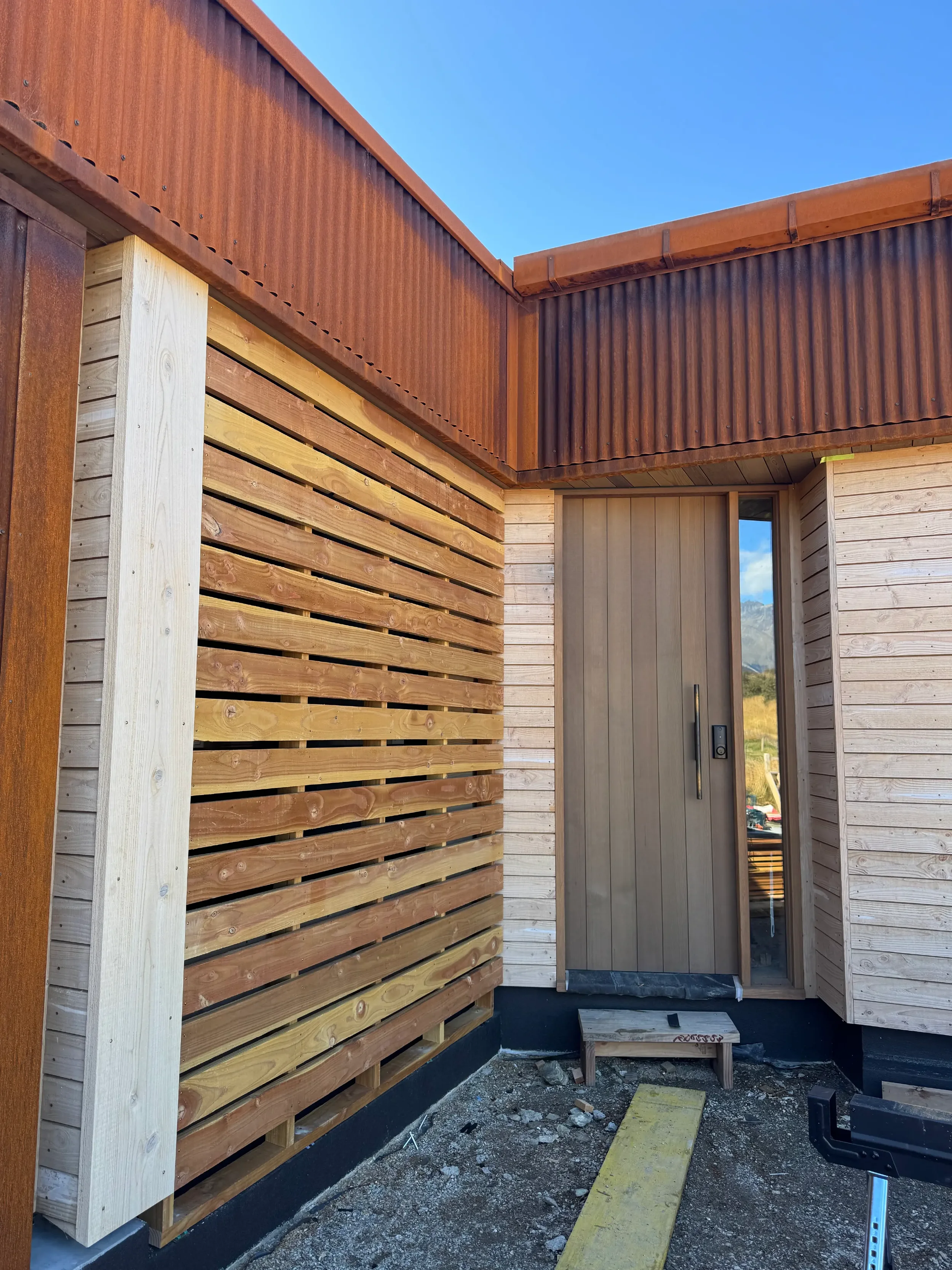 Side of a building under construction with wooden and corrugated metal siding, a door, and a small stepped platform. The sky is blue and mountainous landscape is visible in the distance.