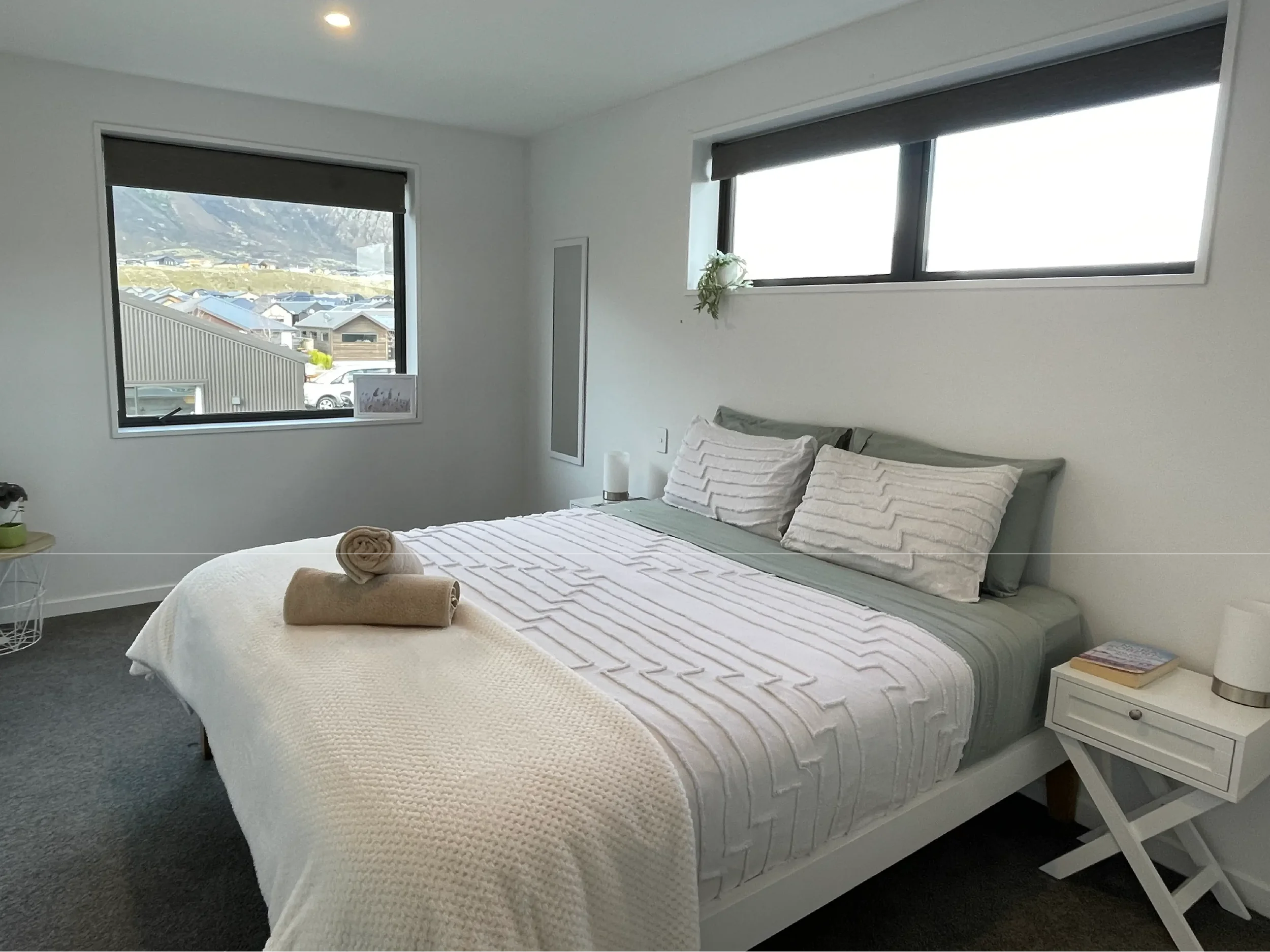 A bedroom with white walls and a bed with white textured bedding and pillows, two beige rolled towels on the bed, a white nightstand with a small book and a lamp, and two large rectangular windows showing an outdoor view of mountains, houses, and par