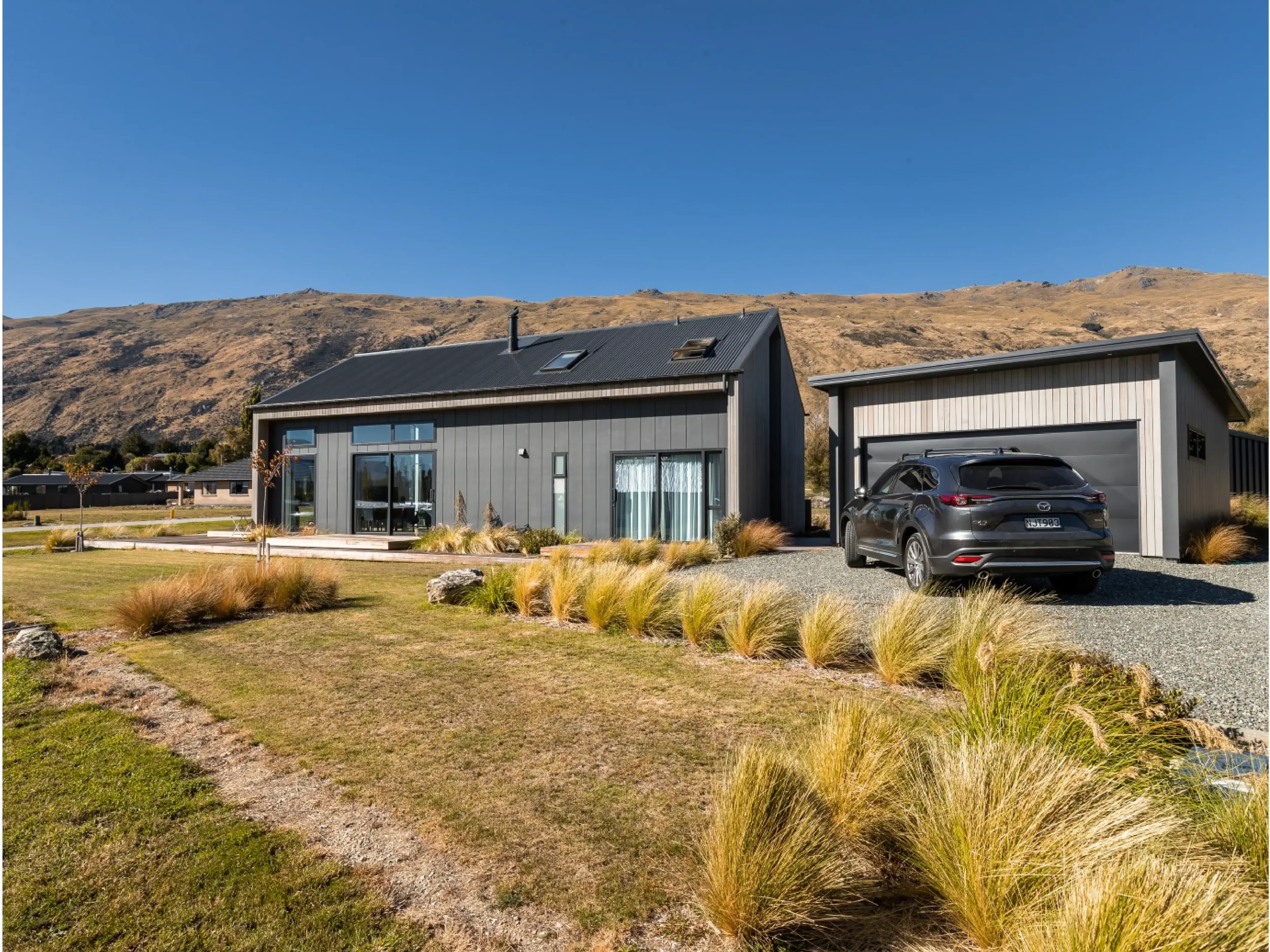 Modern two-story house with gray exterior, large windows, and a black SUV parked in the driveway, surrounded by landscaped grass and plants, with hills in the background under a clear blue sky.