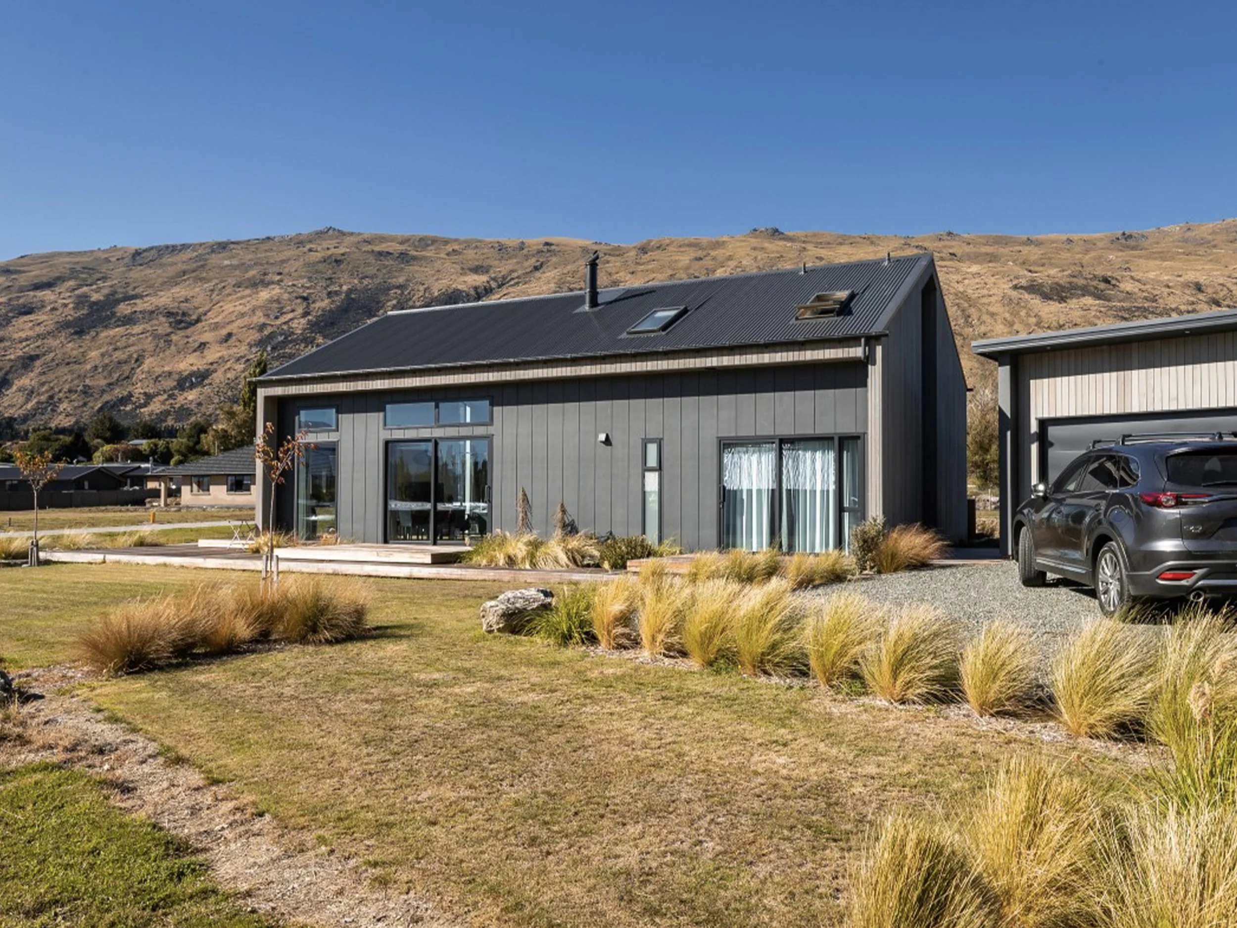 A modern house with large windows and a metal roof, surrounded by a lawn with ornamental grass, with a mountain hill in the background and a blue sky.