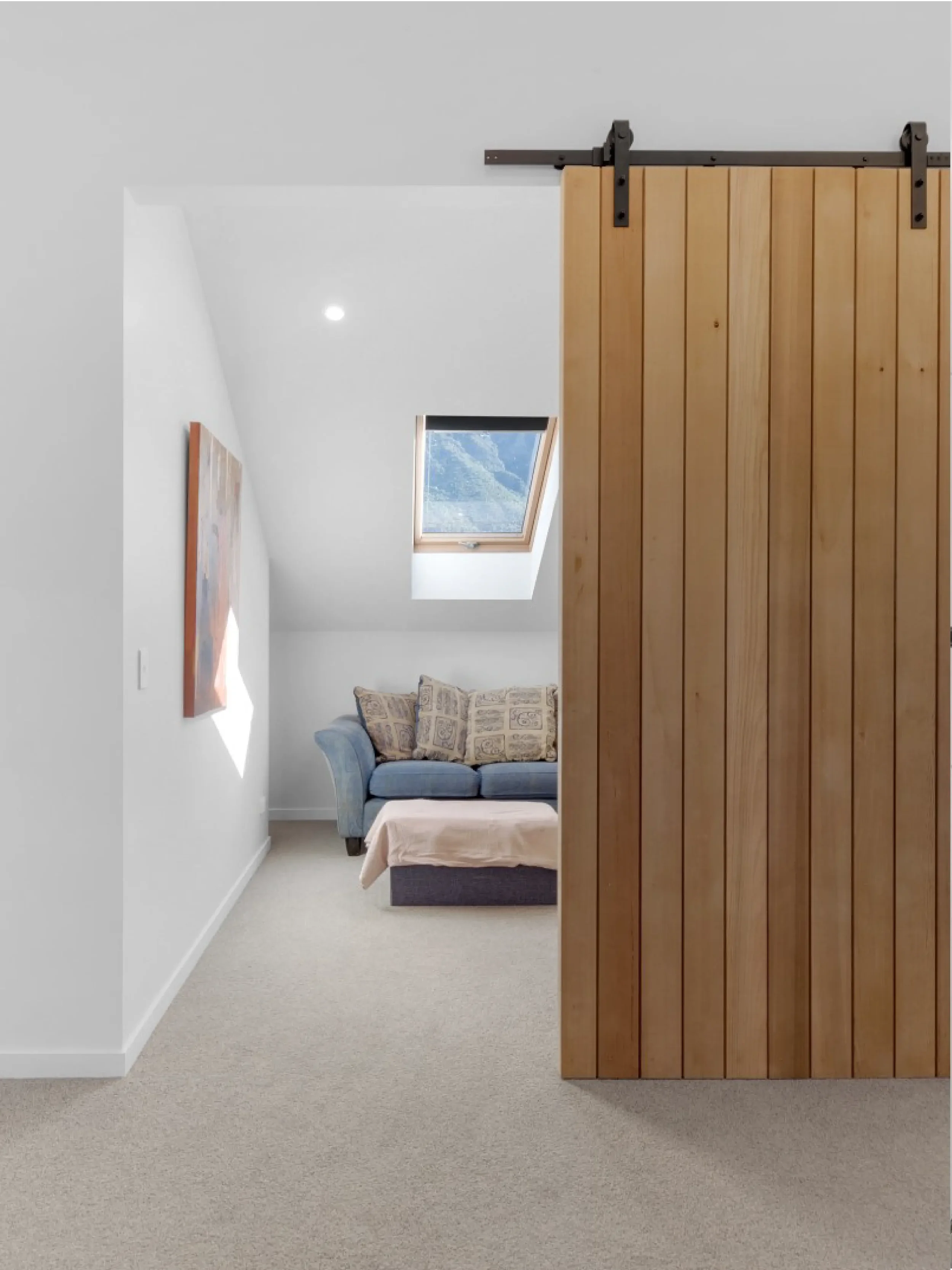A room divided by a wooden barn door on a black metal track, with a sitting area visible behind the door including a blue armchair, pillows, a small table with a cloth, a window with a mountain view, and artwork on the white wall. The room has beige 