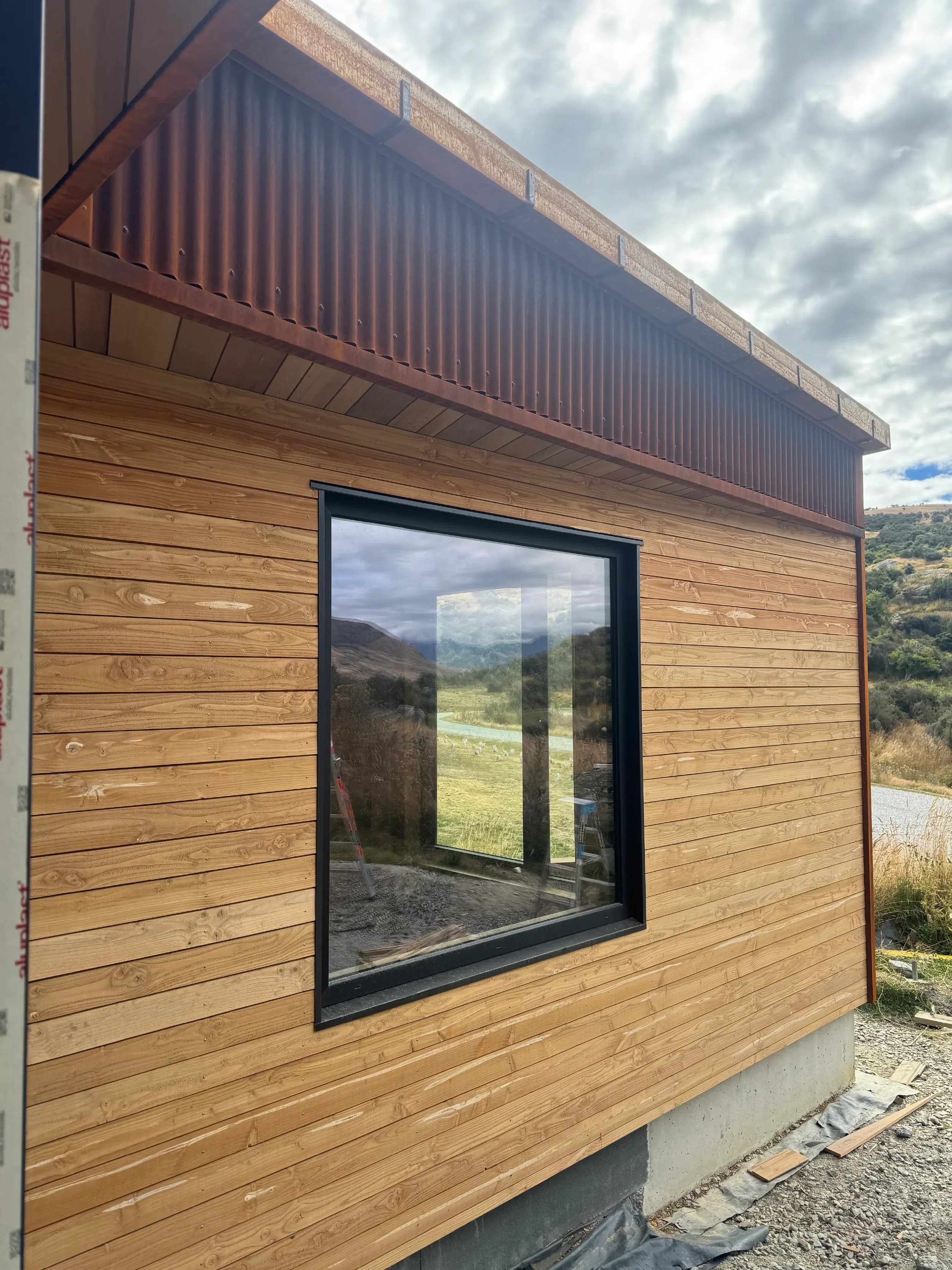 A house under construction with wooden exterior siding, a large window reflecting a scenic landscape, and a cloudy sky above. Construction materials and tools are visible near the base of the house.