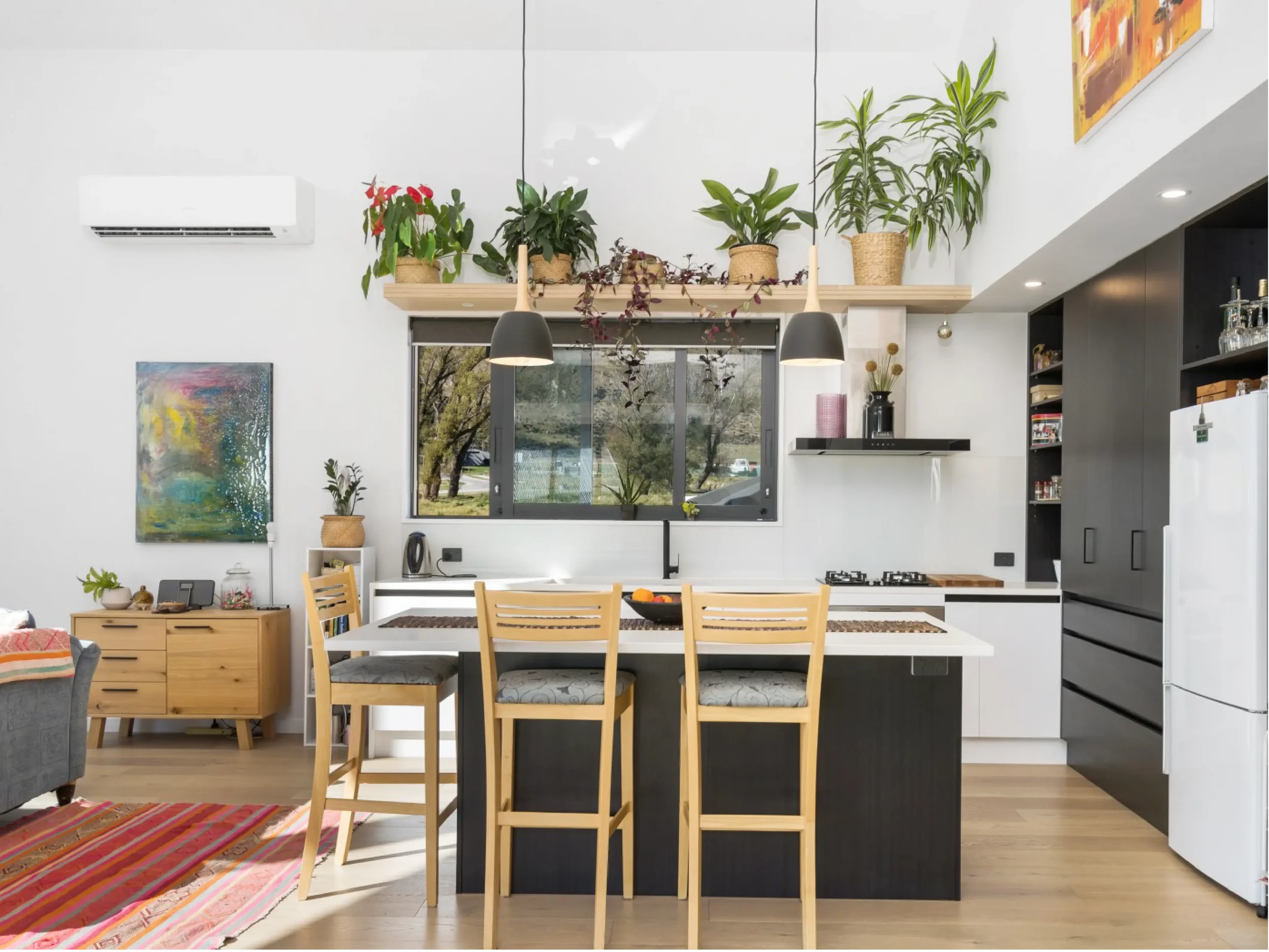 Open kitchen and dining area with a wooden dining table and six chairs, a colorful abstract painting on the wall, a row of potted plants on a high shelf above the window, and a black kitchen cabinet with open shelves.