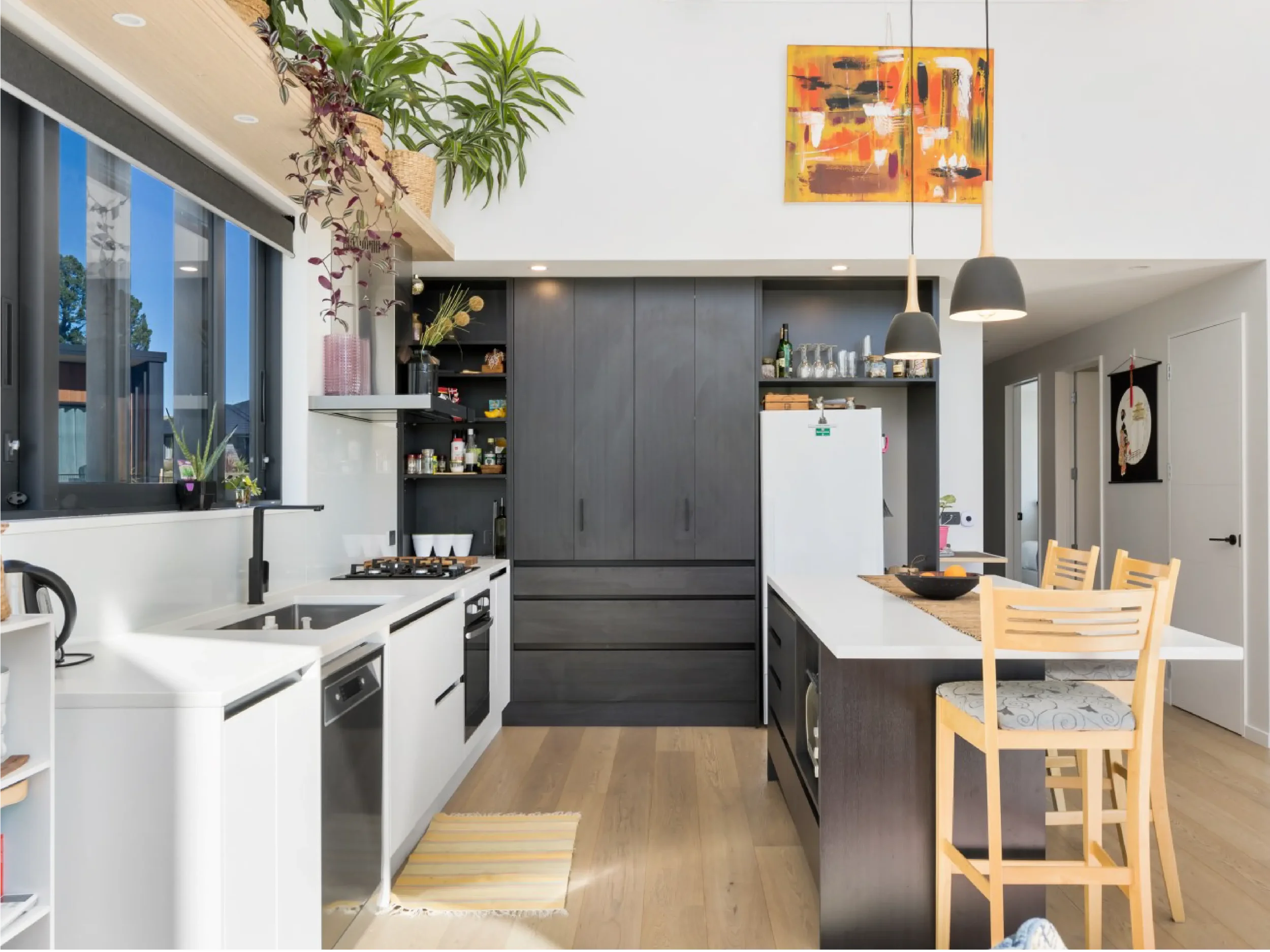 A modern kitchen with white countertops, black cabinets, and hanging black pendant lights. There are plants on a wooden ledge near a large window, with colorful abstract paintings, a small dining table with wooden chairs, and various kitchen items an