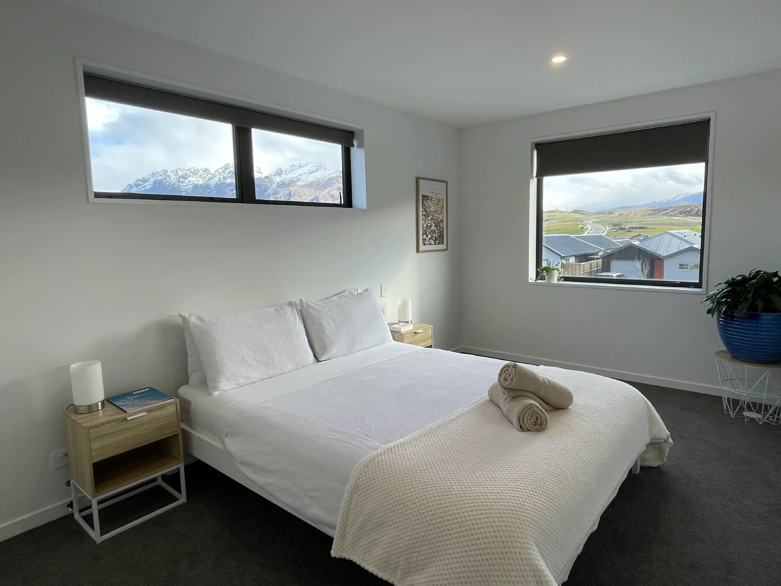 A bedroom with two windows showing snow-capped mountains and green countryside. White bed with pillows, beige blanket, and two rolled towels on top. Nightstand with a lamp and books, framed art on wall, and a potted plant in a blue pot.