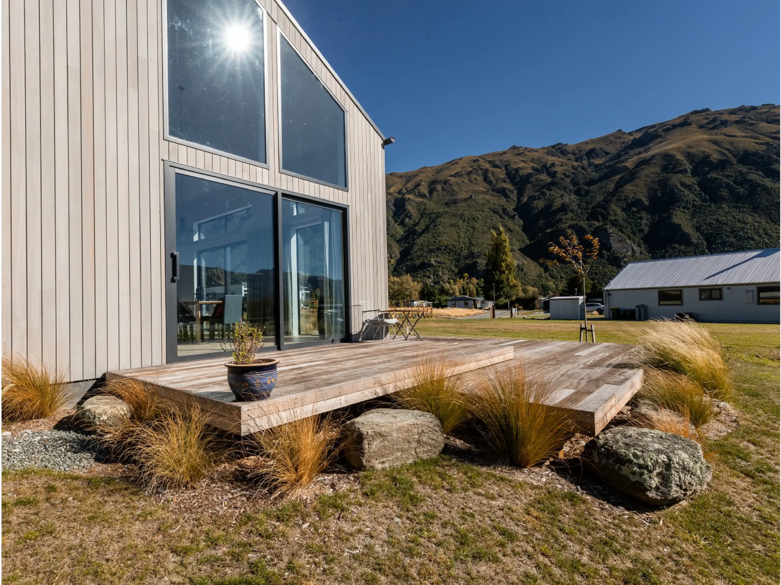 A modern building with large glass windows, situated in a mountainous landscape with clear blue sky. There is a wooden deck with outdoor chairs and potted plants in the foreground, and other small buildings and trees nearby.