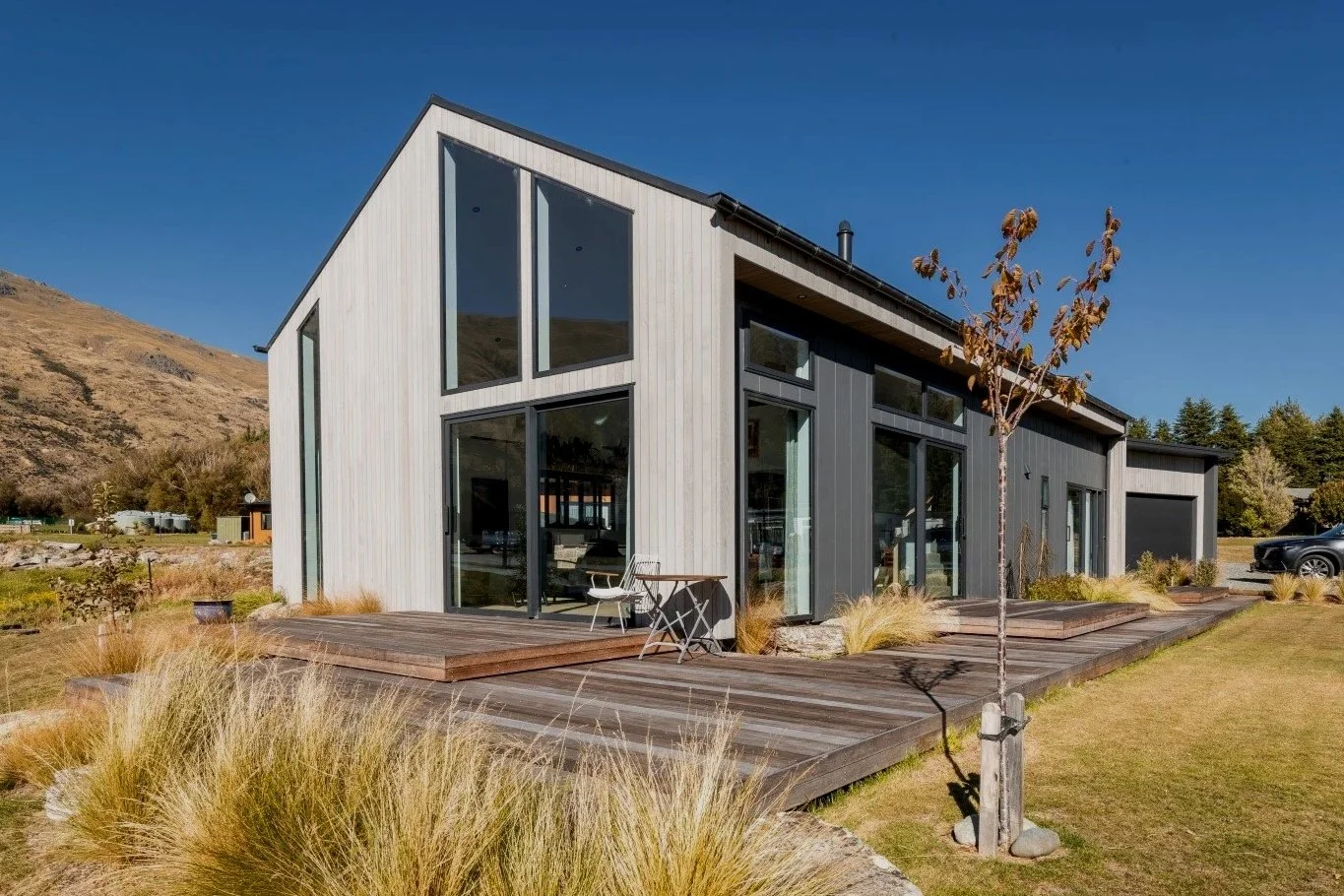 Modern house with large glass windows and a wooden deck in a natural landscape with hills and sparse trees.