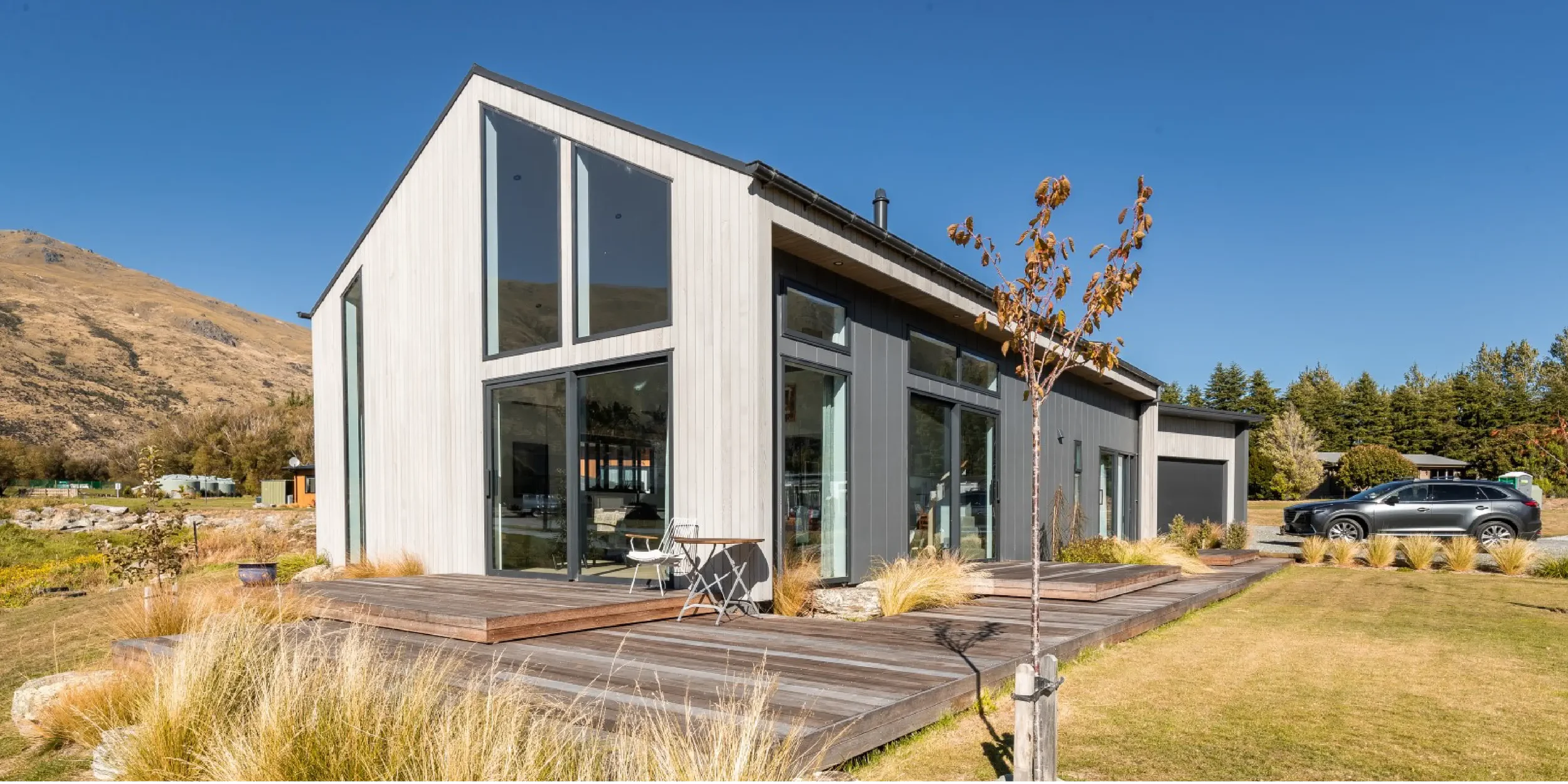Modern two-story house with large windows, wooden deck, and gray exterior, set against a mountain landscape with clear blue sky.