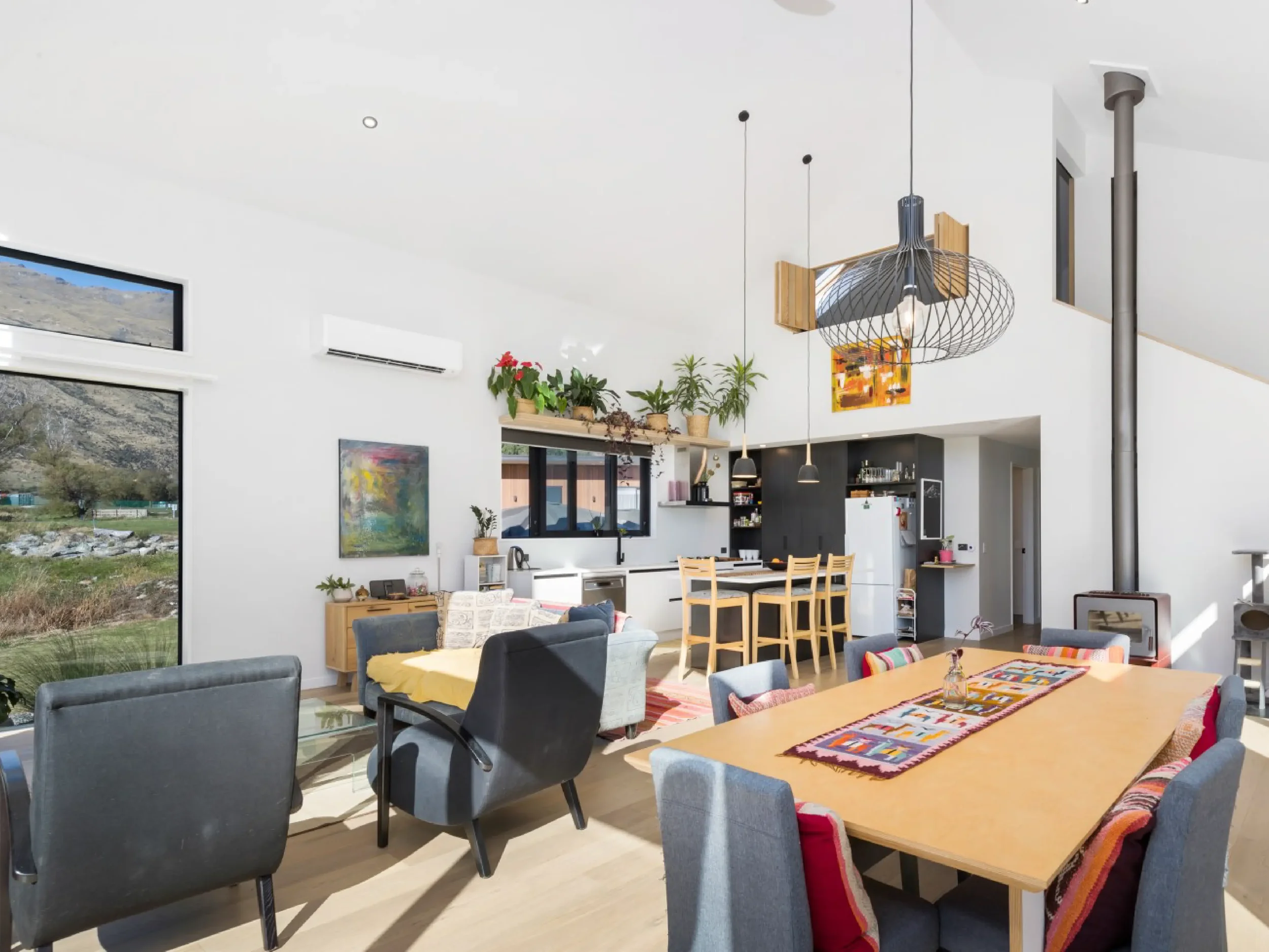 Interior of a modern, bright living and dining room with large windows, with a kitchen area in the background. The space features dining tables, cushioned chairs, and decorative plants on a shelf, with hanging pendant lights and artistic wall decor.