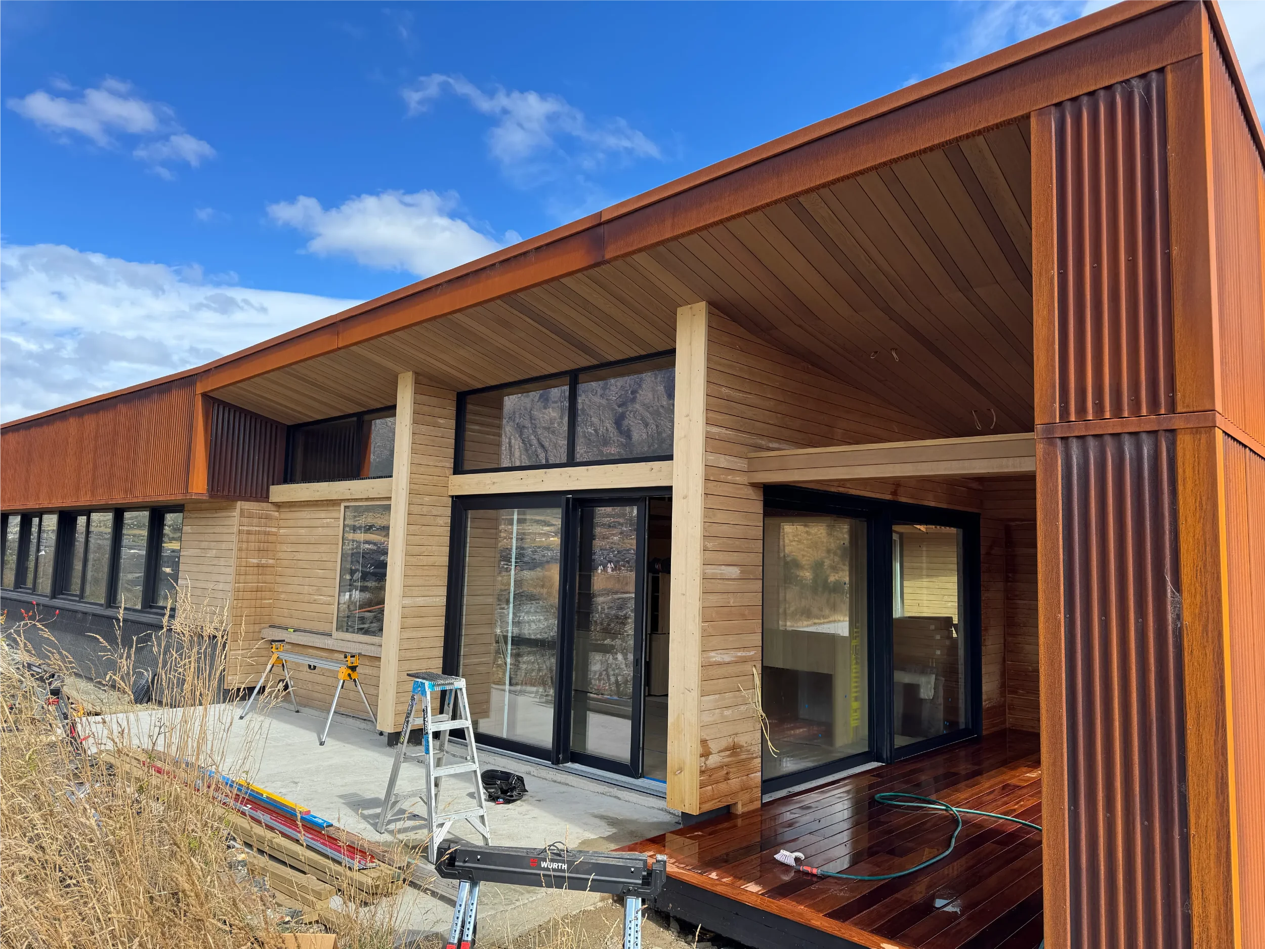 Modern house with wooden exterior, large glass windows, and a flat roof, under a blue sky with scattered clouds. Construction tools and materials are visible in the foreground, indicating ongoing construction.