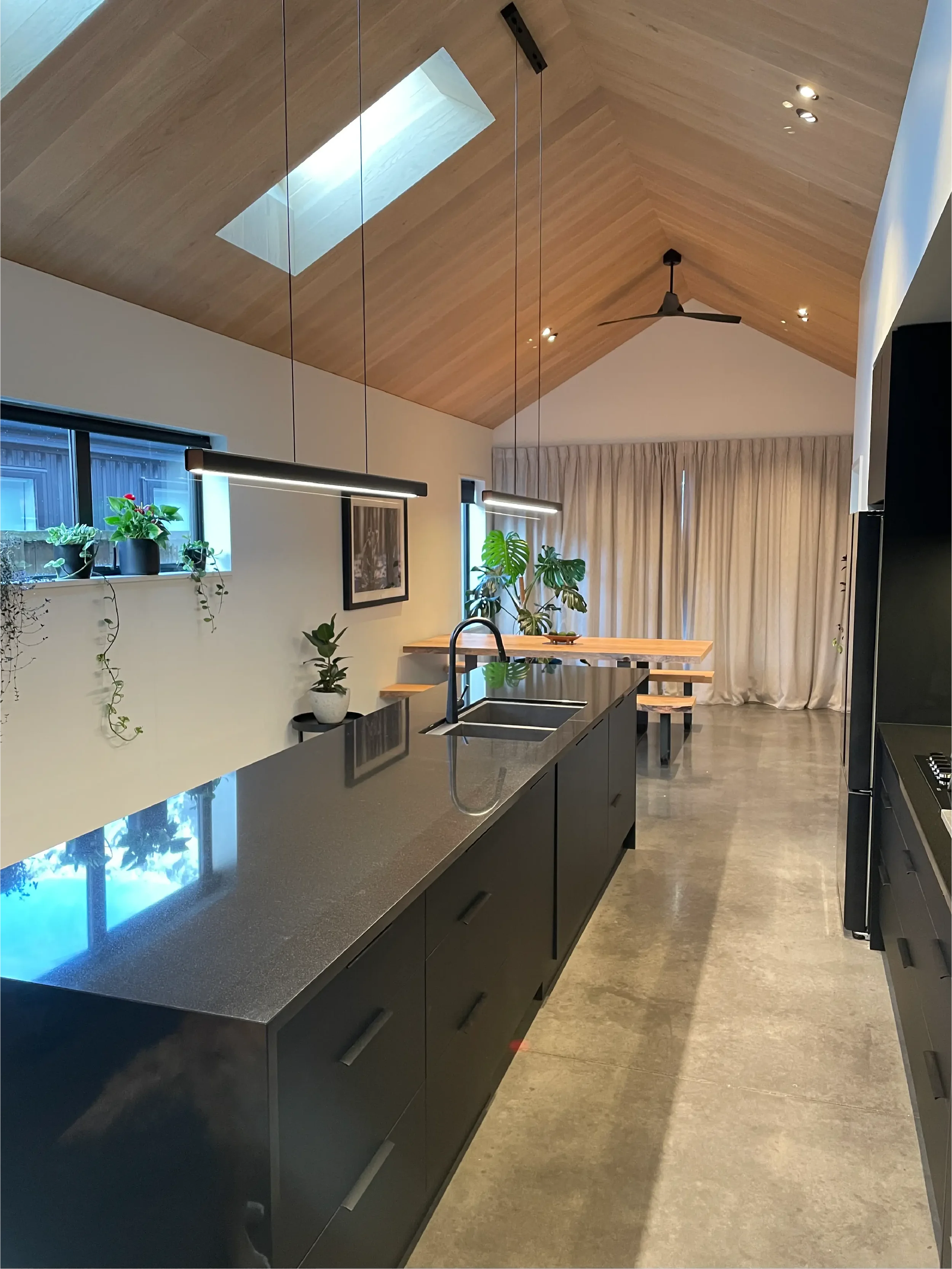 Modern kitchen with black cabinetry, a black island with a sink, hanging pendant lights, and indoor plants near a window and a wooden dining table.