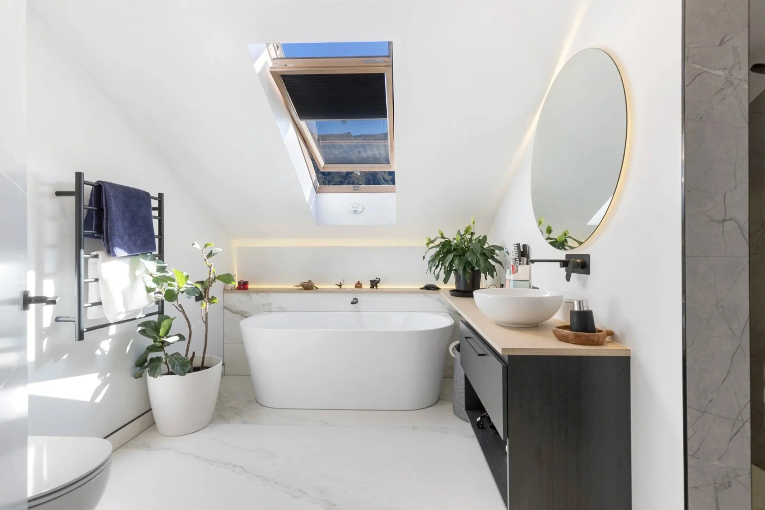 Modern bathroom with a freestanding bathtub, a large round mirror with backlit lighting, a vessel sink with a black faucet, and a large window with a view of mountains. There are green plants and decorative items on the counter and shelf, with a towel rack on the wall.