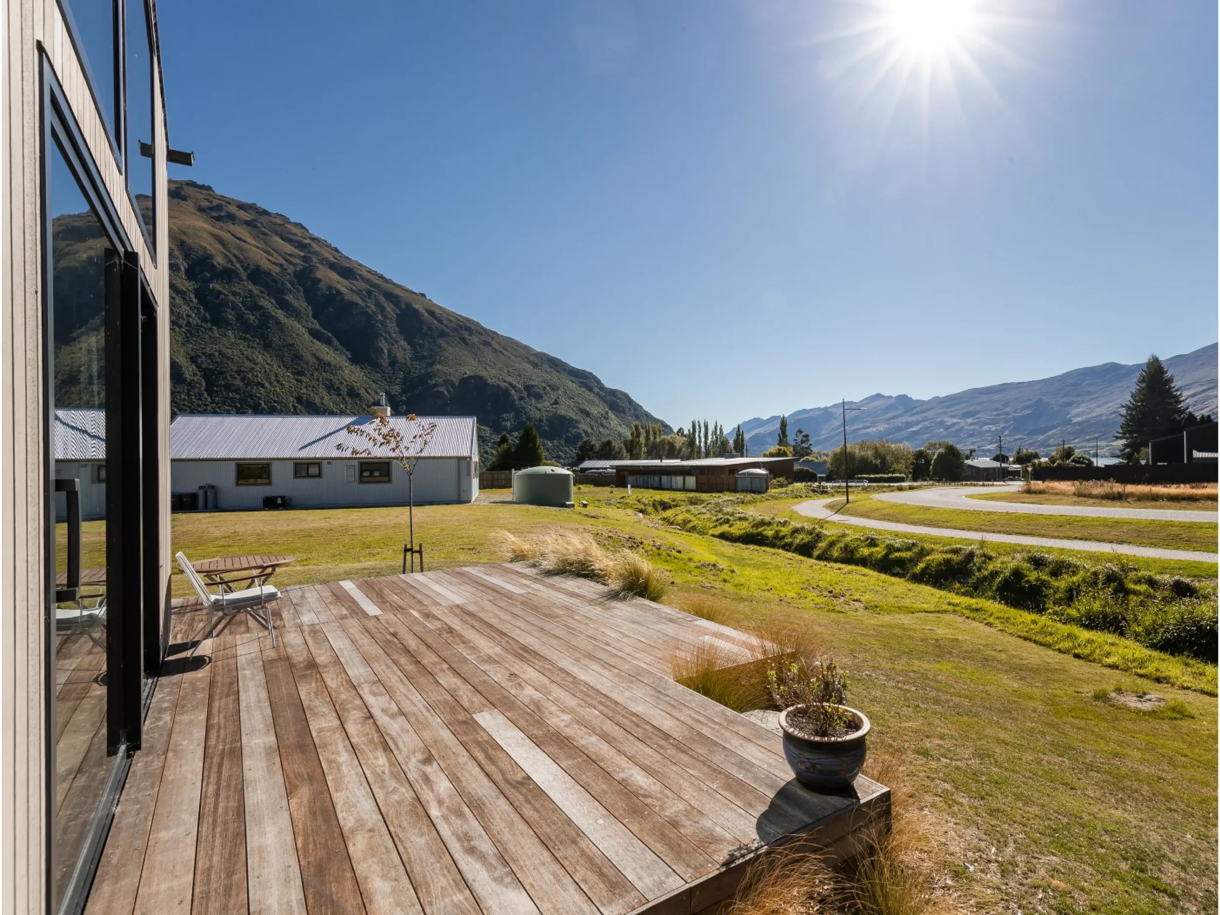 View from a house deck overlooking a grassy yard with trees and buildings, mountain landscape in the background, bright sun shining in a clear blue sky.
