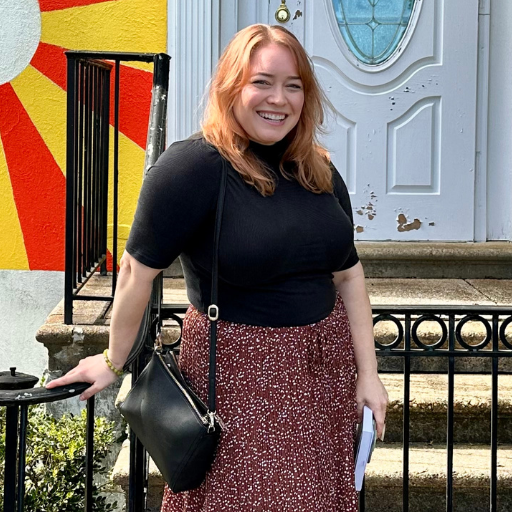 A smiling woman with red hair standing on stairs in front of a colorful mural and a blue door, holding a book.