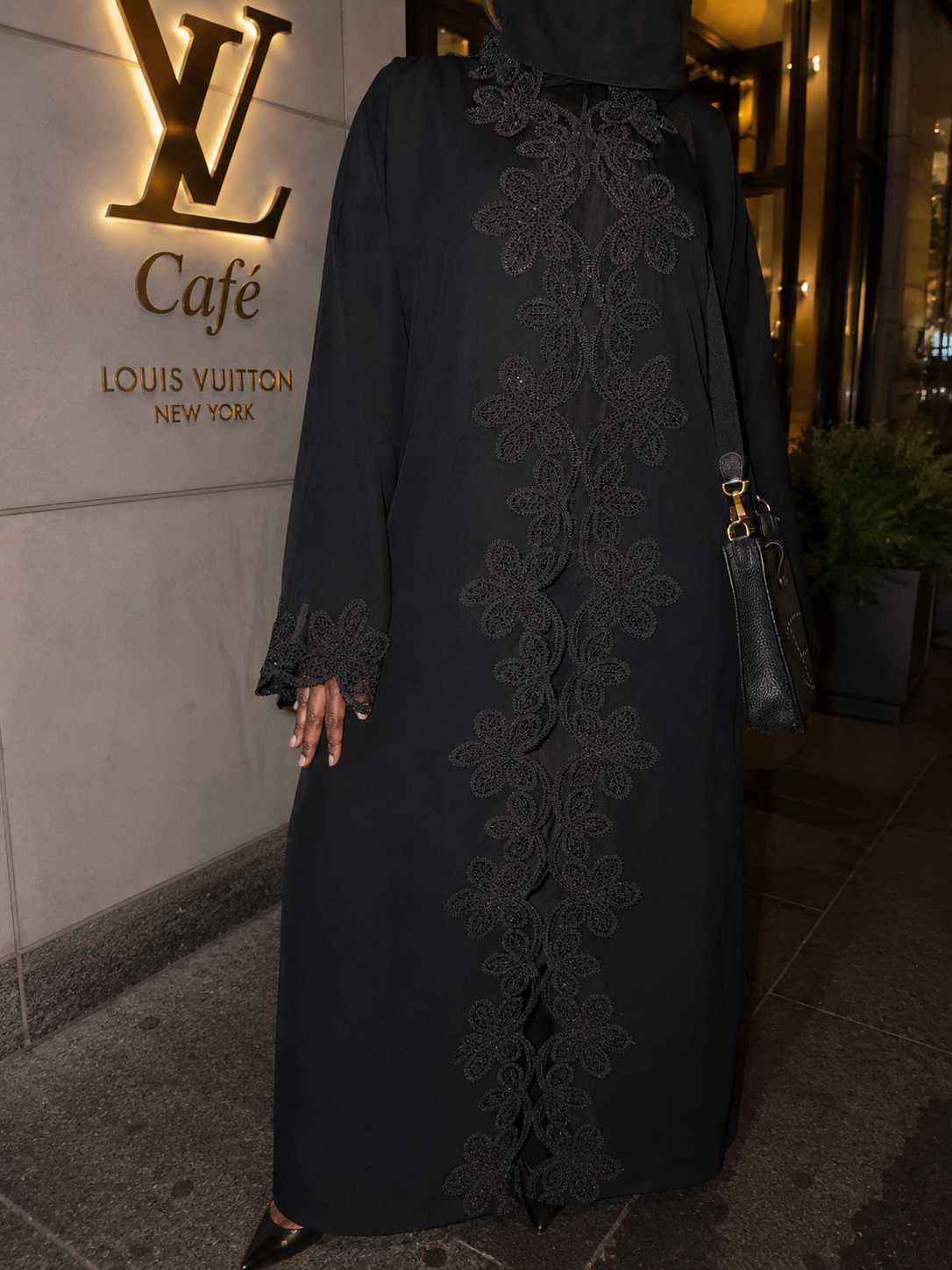 Woman wearing a long black abaya with intricate lace embroidery standing outside Louis Vuitton Café, New York.