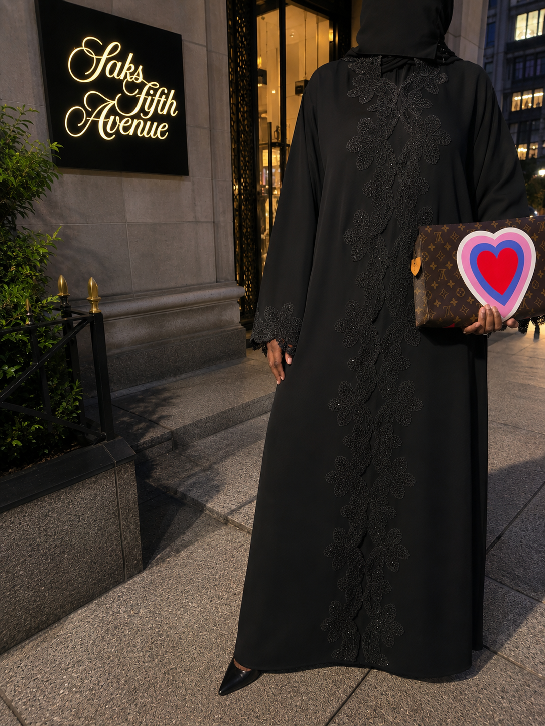 A woman dressed in a long black abaya, holding a Louis Vuitton toiletry 25 bag with a colorful heart sticker, stands in front of Saks Fifth Avenue at dusk. The store's illuminated sign is visible on the left.