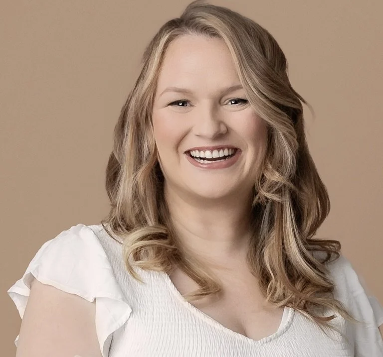 A smiling woman with wavy blonde hair, wearing a white top with ruffled sleeves, standing against a plain beige background.